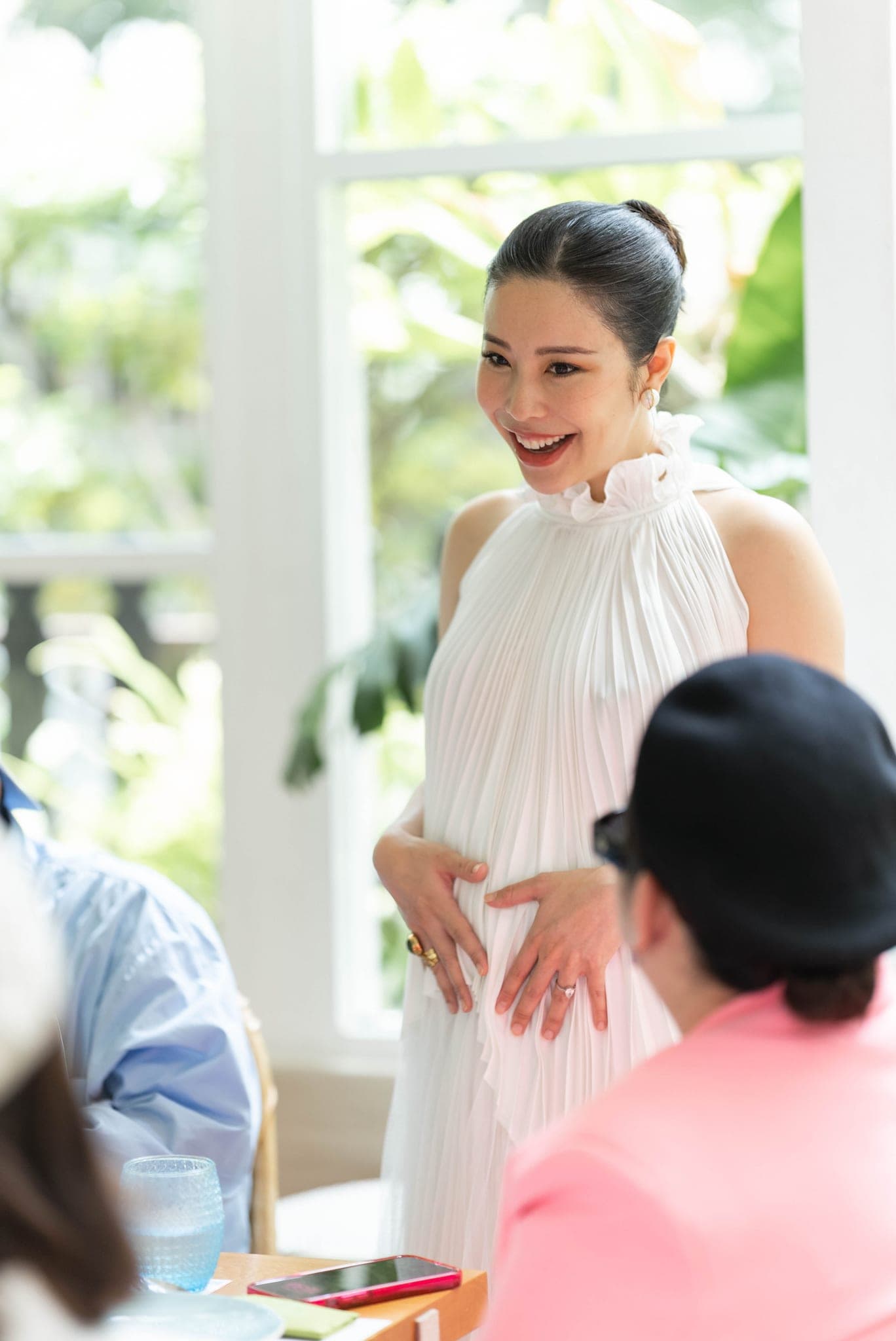 Hands on her bump, laughing down at a friend at the table
