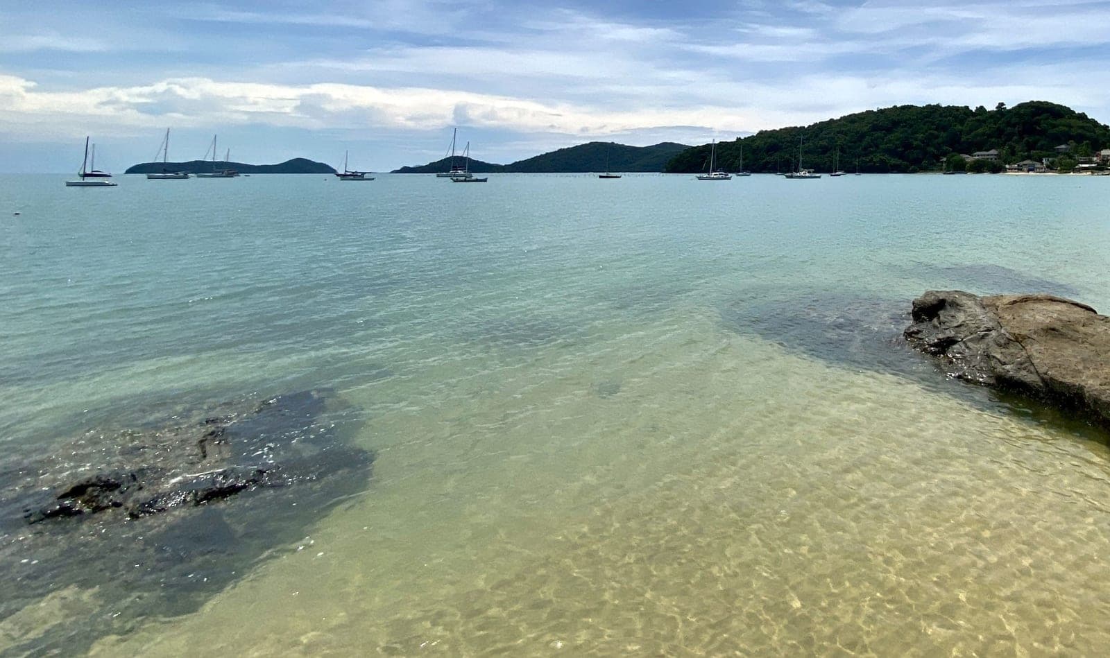 Clear, calm shallows over pale sand and dark rocks at Ao Yon, yachts at anchor across the bay and green hills behind