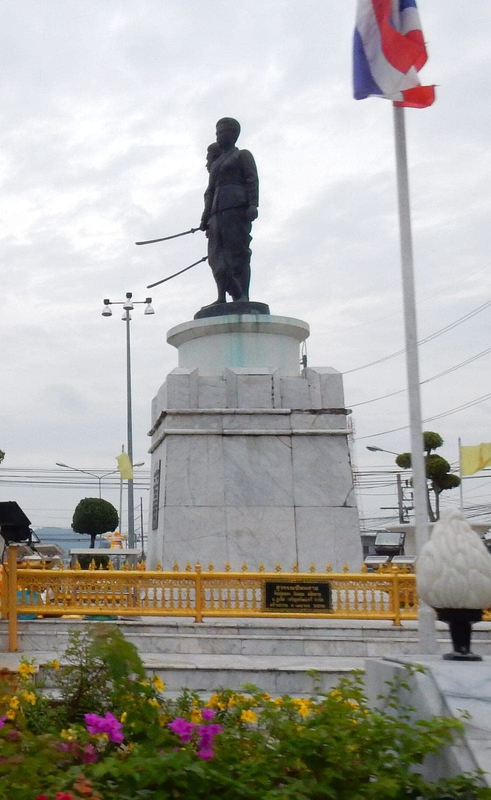 Two bronze women with swords on a tall white plinth behind a gold railing, a Thai flag and flower beds around them under a grey sky