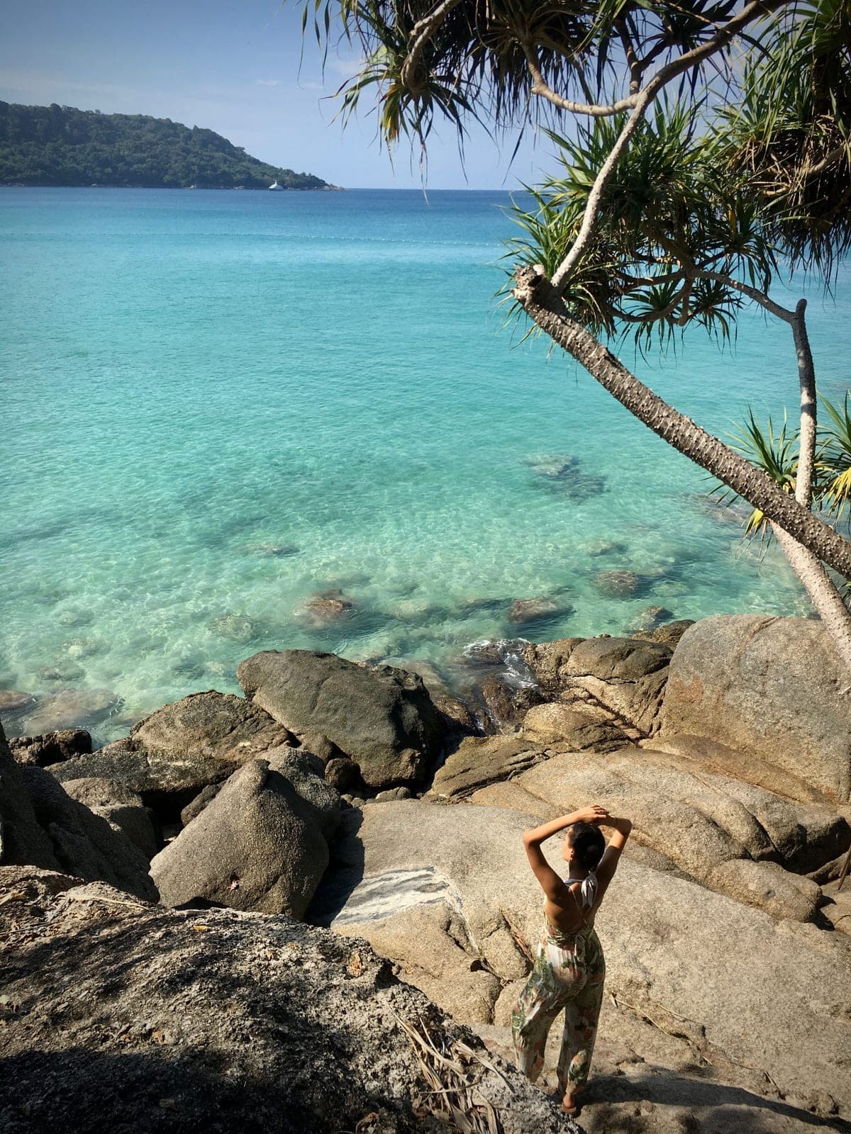 A woman on her own on the granite boulders above clear turquoise water at Karon, her back to the camera, a screw pine leaning over the sea