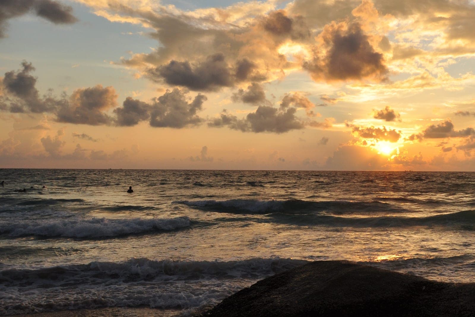 The sun setting under gold and grey clouds, waves breaking on the shore and a dark boulder in the foreground