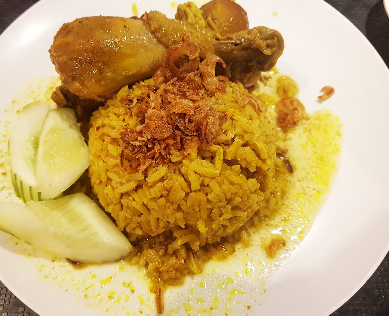Khao mok kai: a mound of yellow rice topped with fried shallots, braised chicken behind it and cucumber beside it on a white plate
