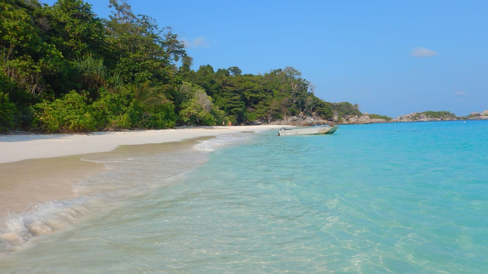 White sand and clear turquoise shallows, jungle running down to the beach and a small boat at anchor