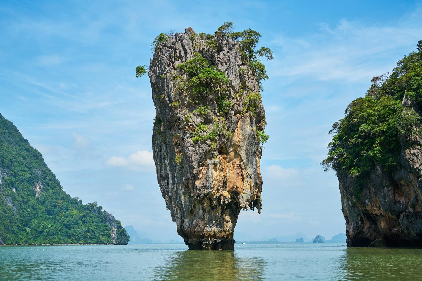 A tall limestone pillar topped with trees standing alone in calm green water, a cliff on either side