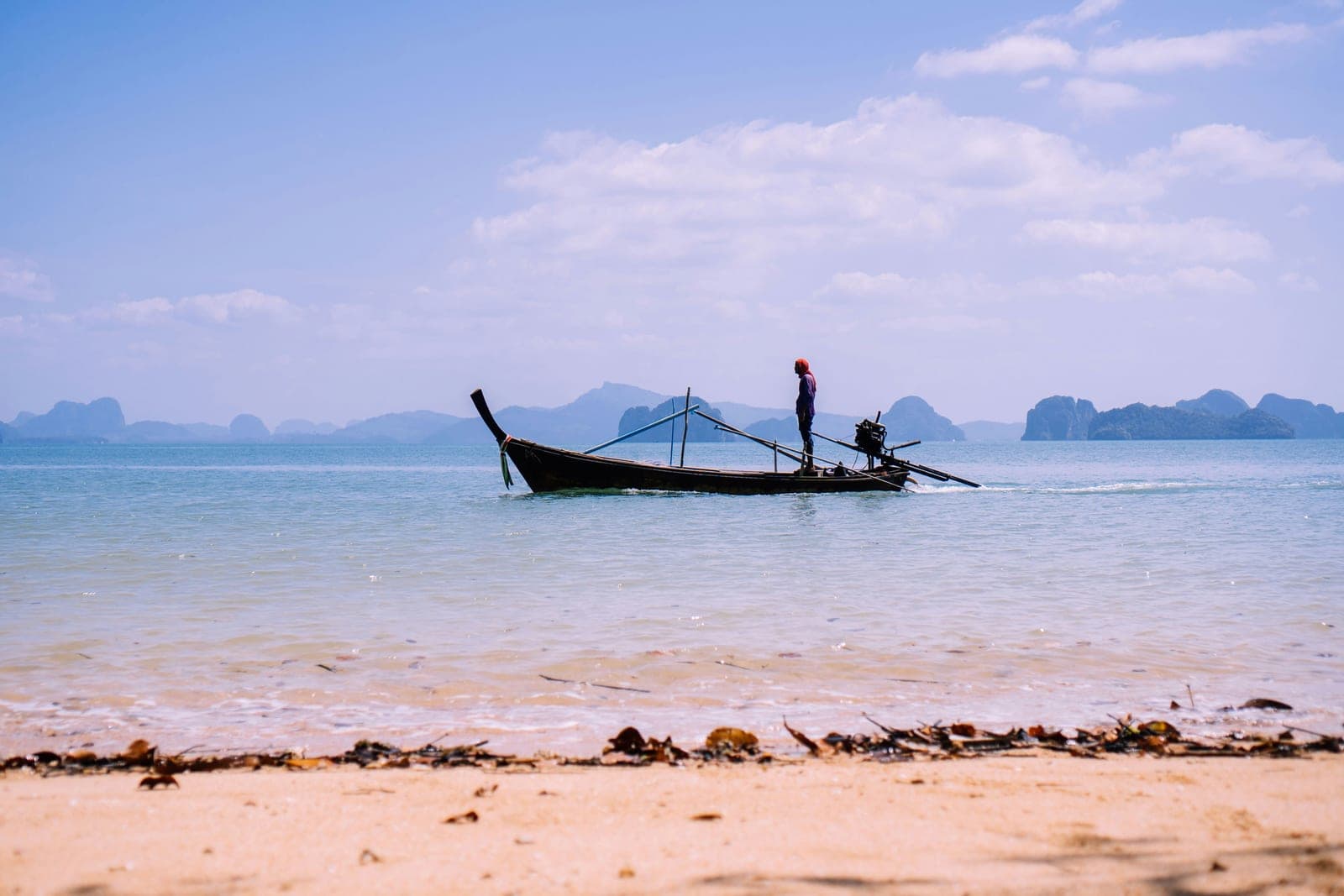 A boatman standing at the stern of a wooden longtail on pale blue water, a row of limestone islands on the horizon