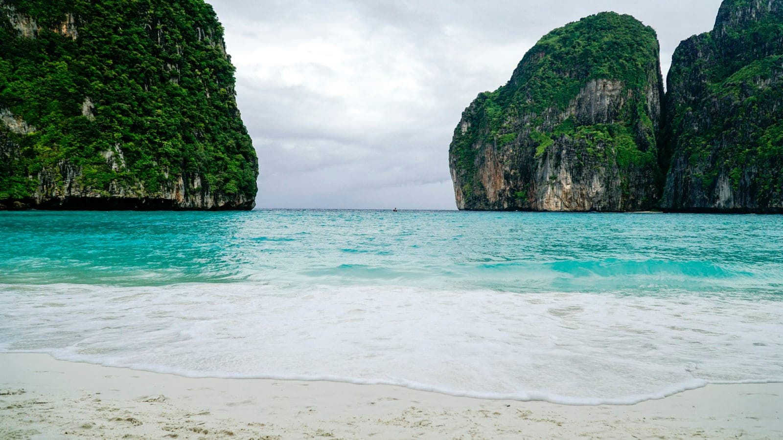 An empty white beach and turquoise water between two jungle-covered cliffs under a grey sky