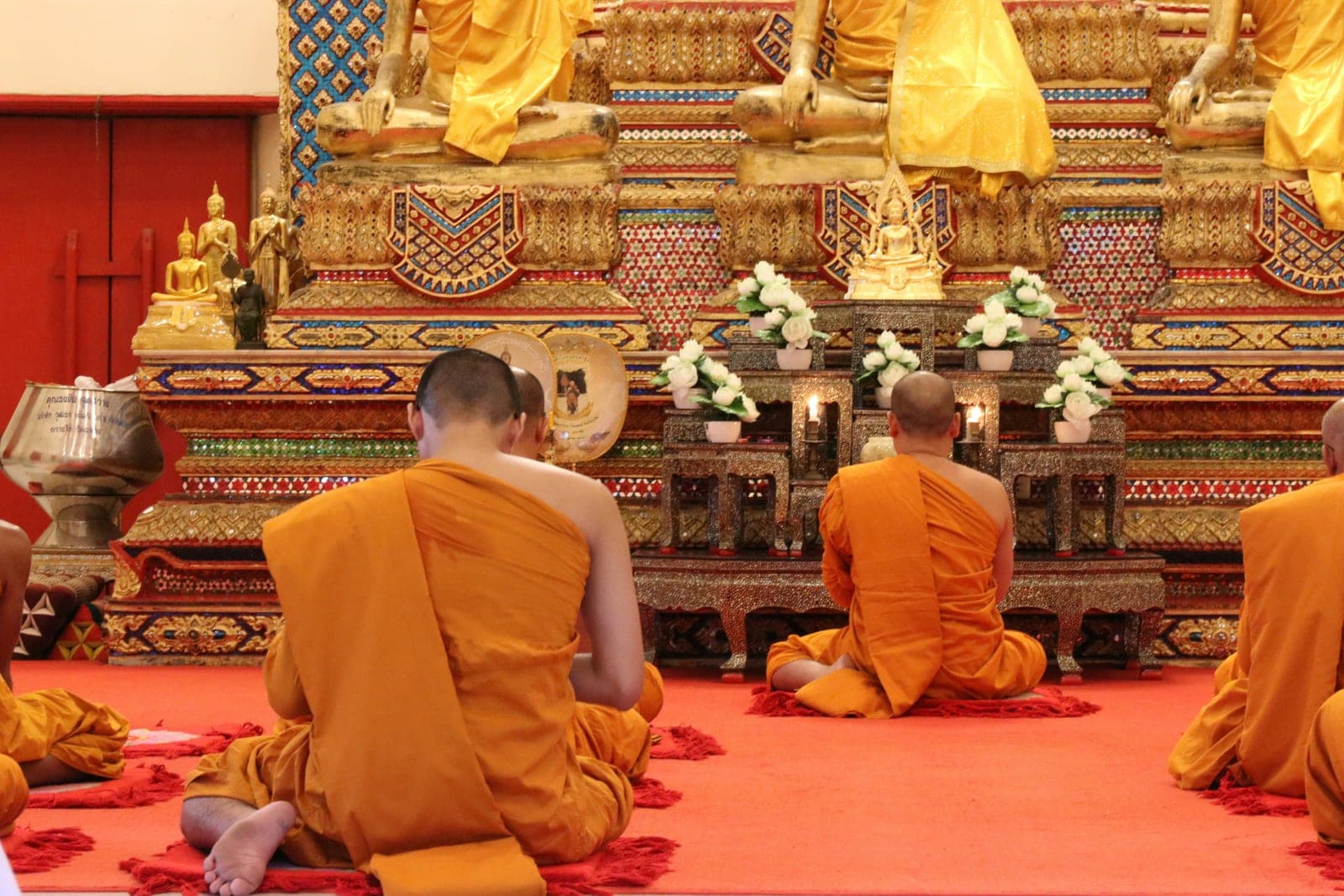 Monks in saffron robes kneeling in prayer, seen from behind, before gilded Buddha images in a temple hall