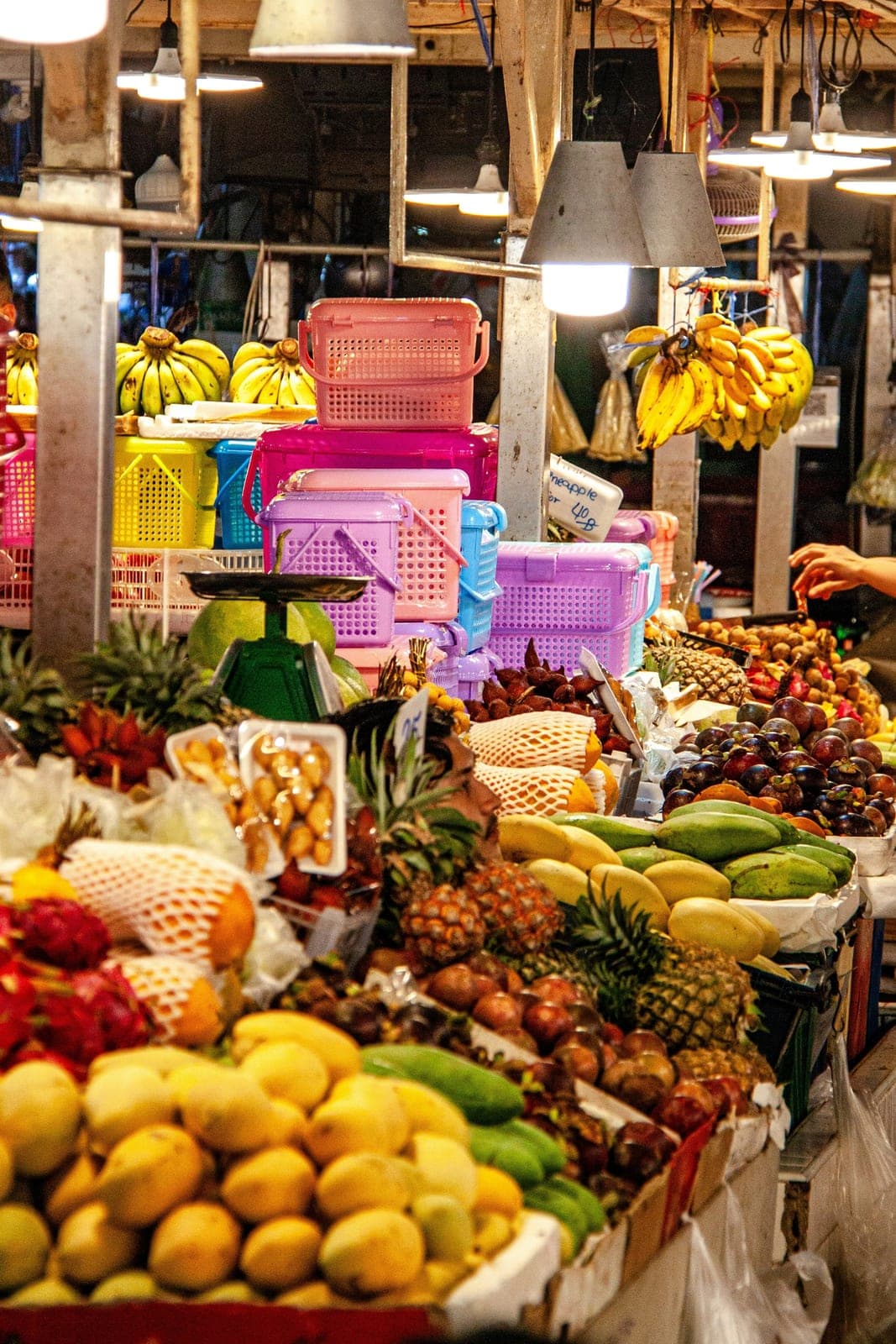 Mangoes, pineapples, mangosteens and bananas piled on a market stall under hanging lamps