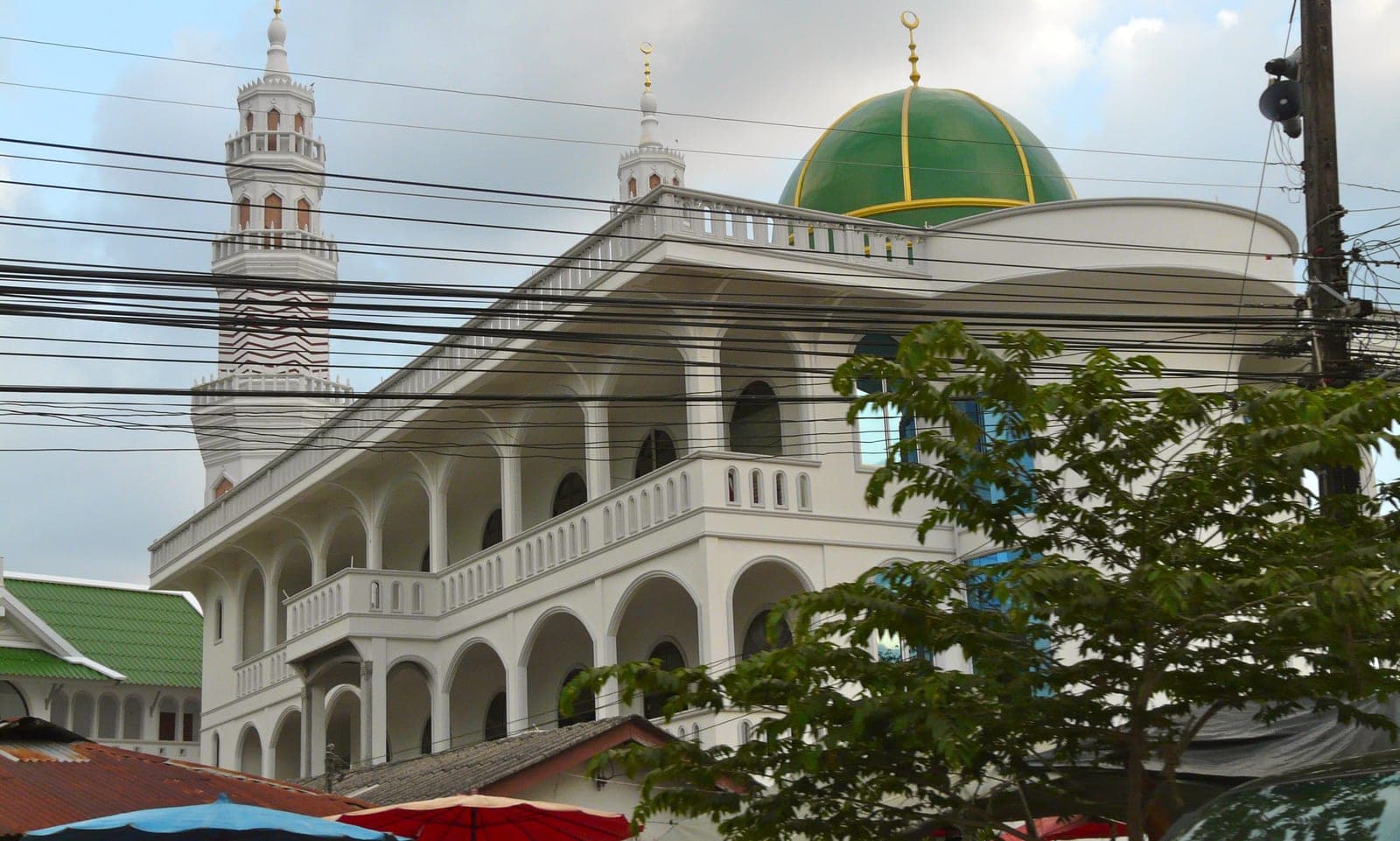 A white mosque with arched balconies, a green dome edged in gold and a tall minaret, power lines across the front and a tree to one side