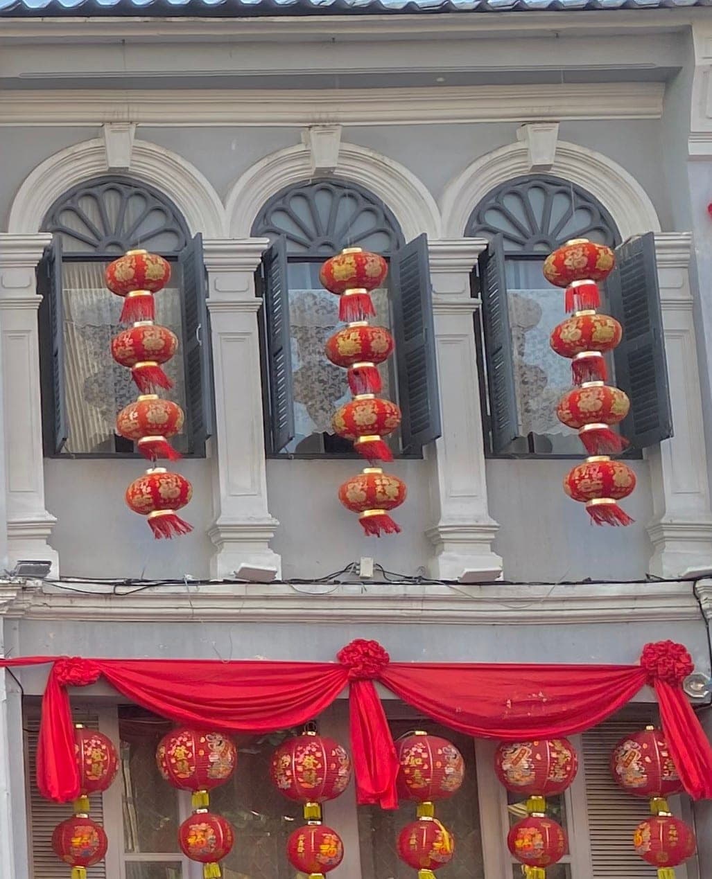 Strings of red lanterns hanging at the arched, shuttered windows of an Old Town shophouse, red swags and more lanterns over the shopfront below