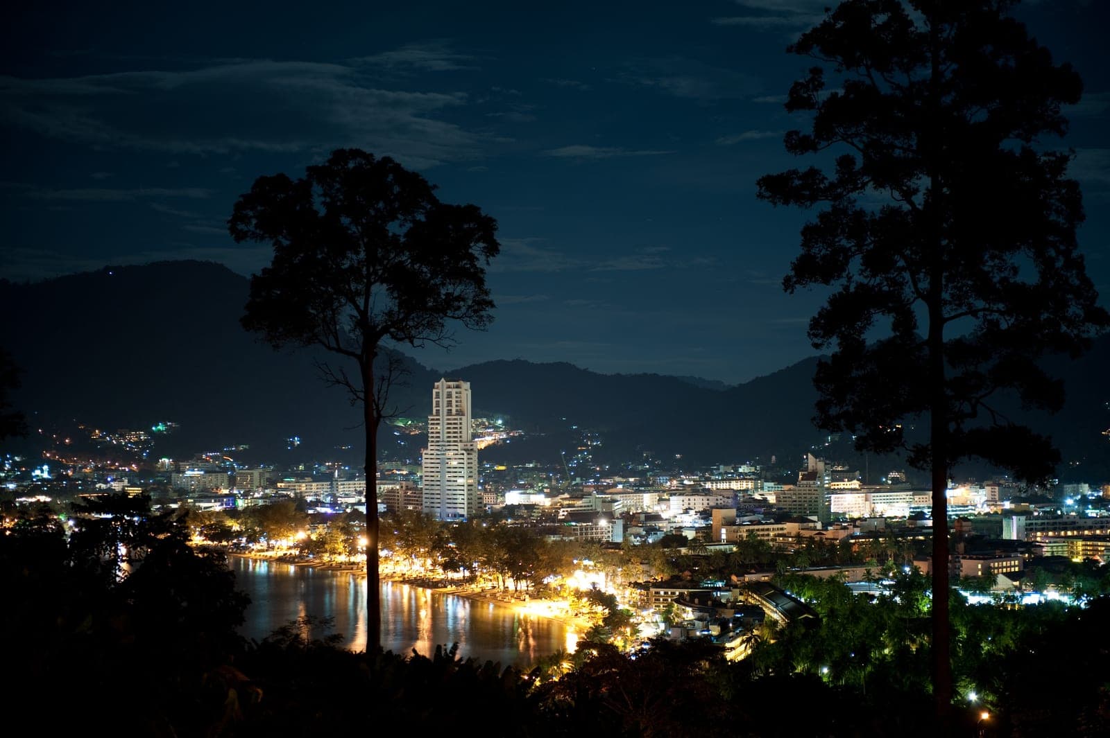 Patong at night from a wooded hillside, the lit curve of the beach road reflected in the bay, a tall tower among the town's lights