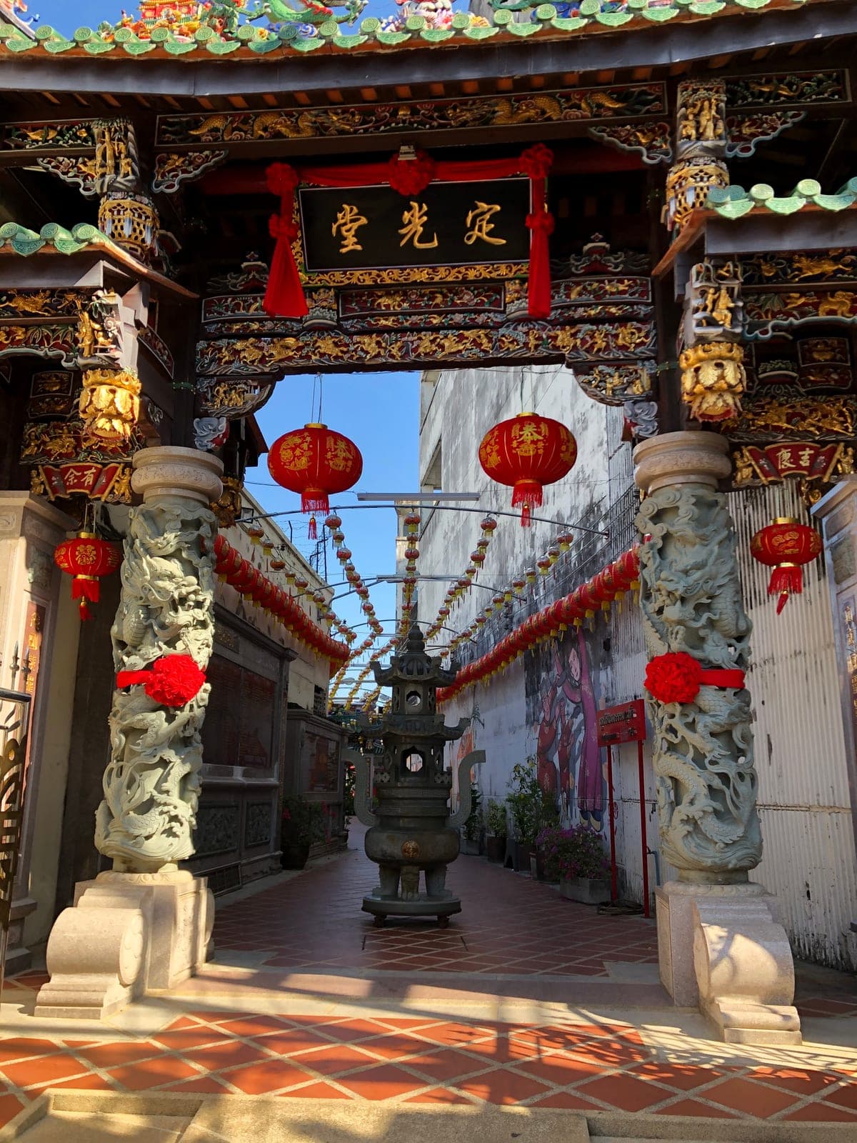 A carved shrine gate hung with red lanterns and ribbons, stone dragon pillars either side and a lane strung with lanterns beyond