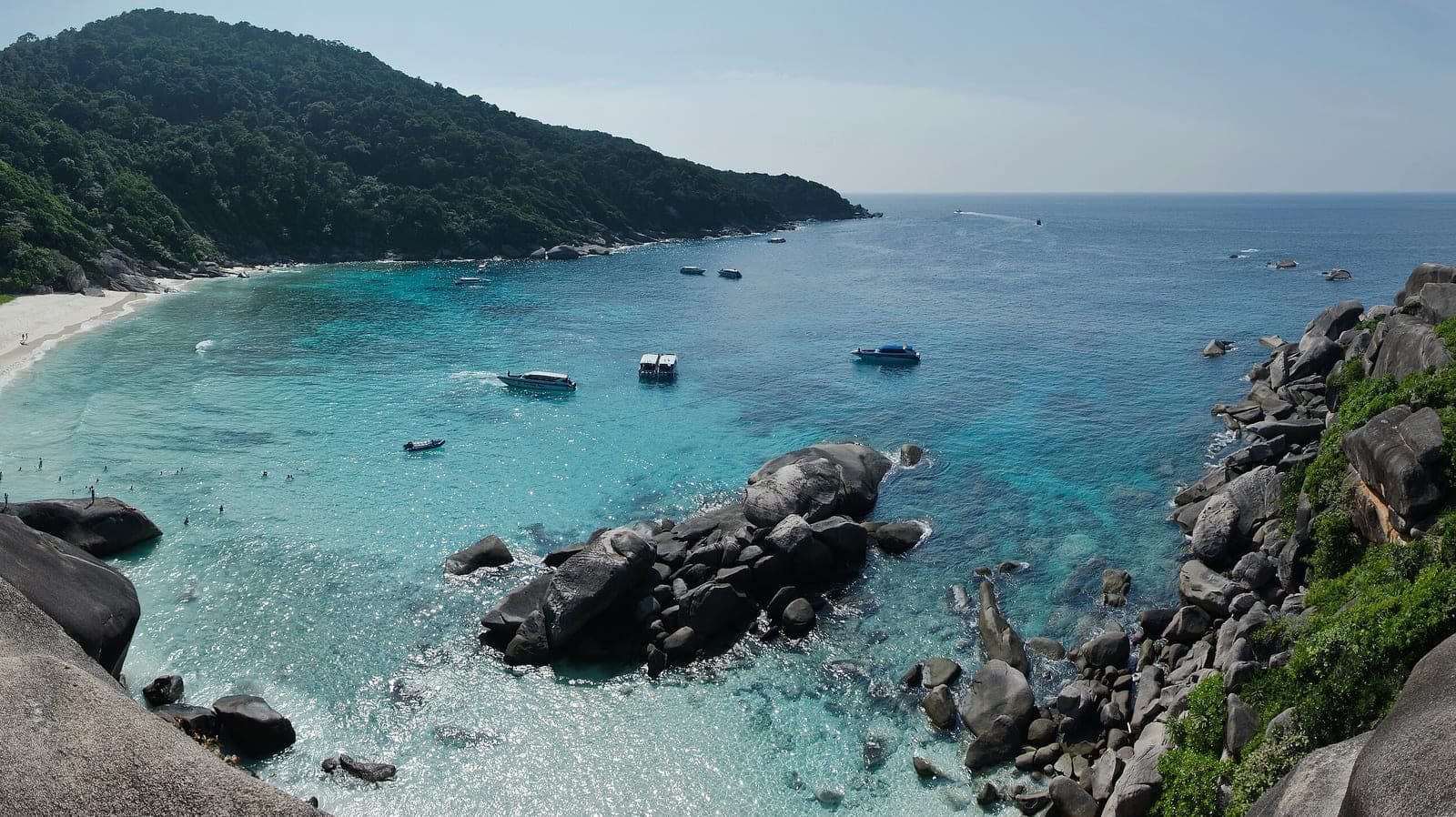 A white beach curving round a turquoise bay below a jungle hill, speedboats at anchor and granite boulders in the foreground