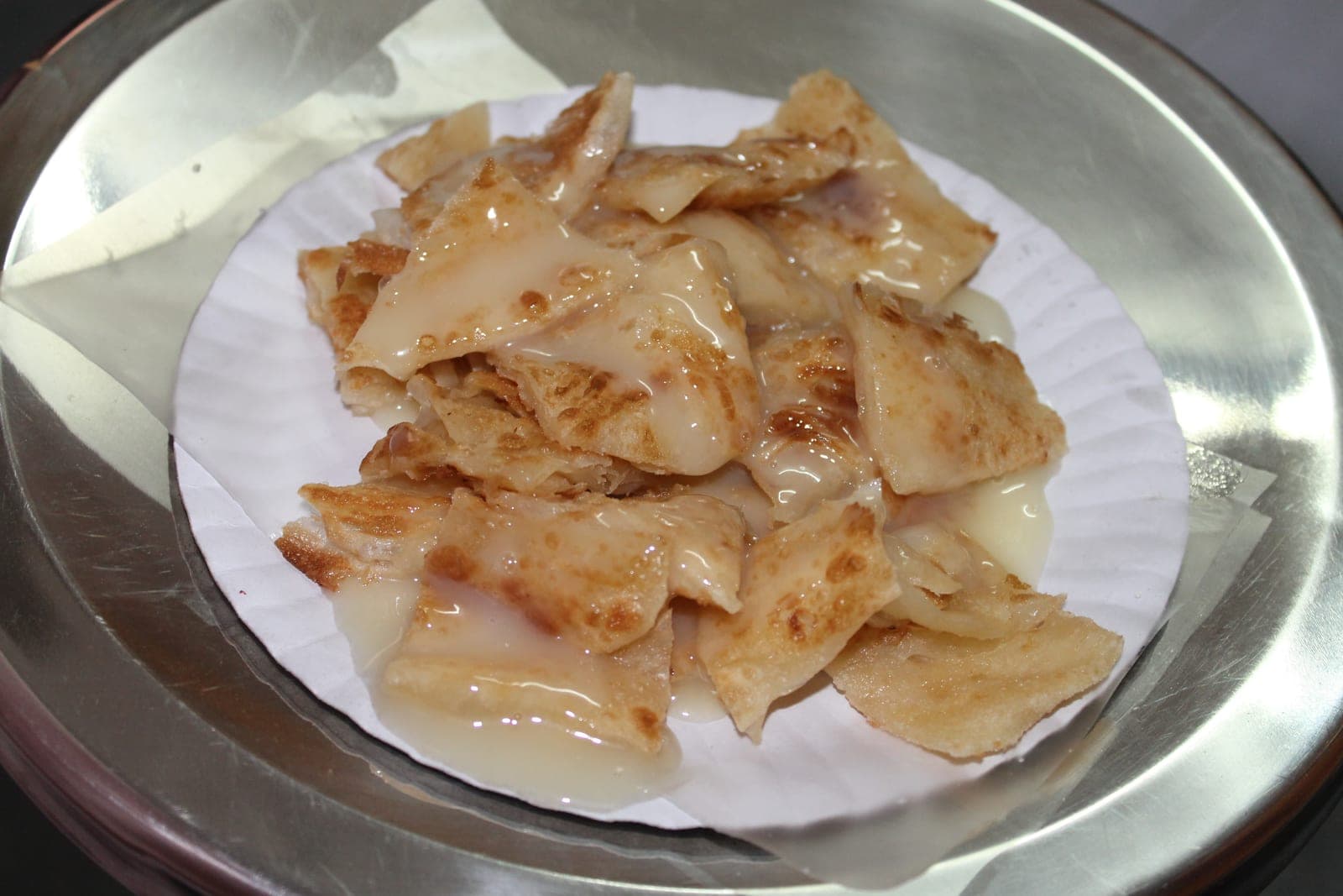 Torn pieces of crisp roti under condensed milk on a paper plate on a steel tray
