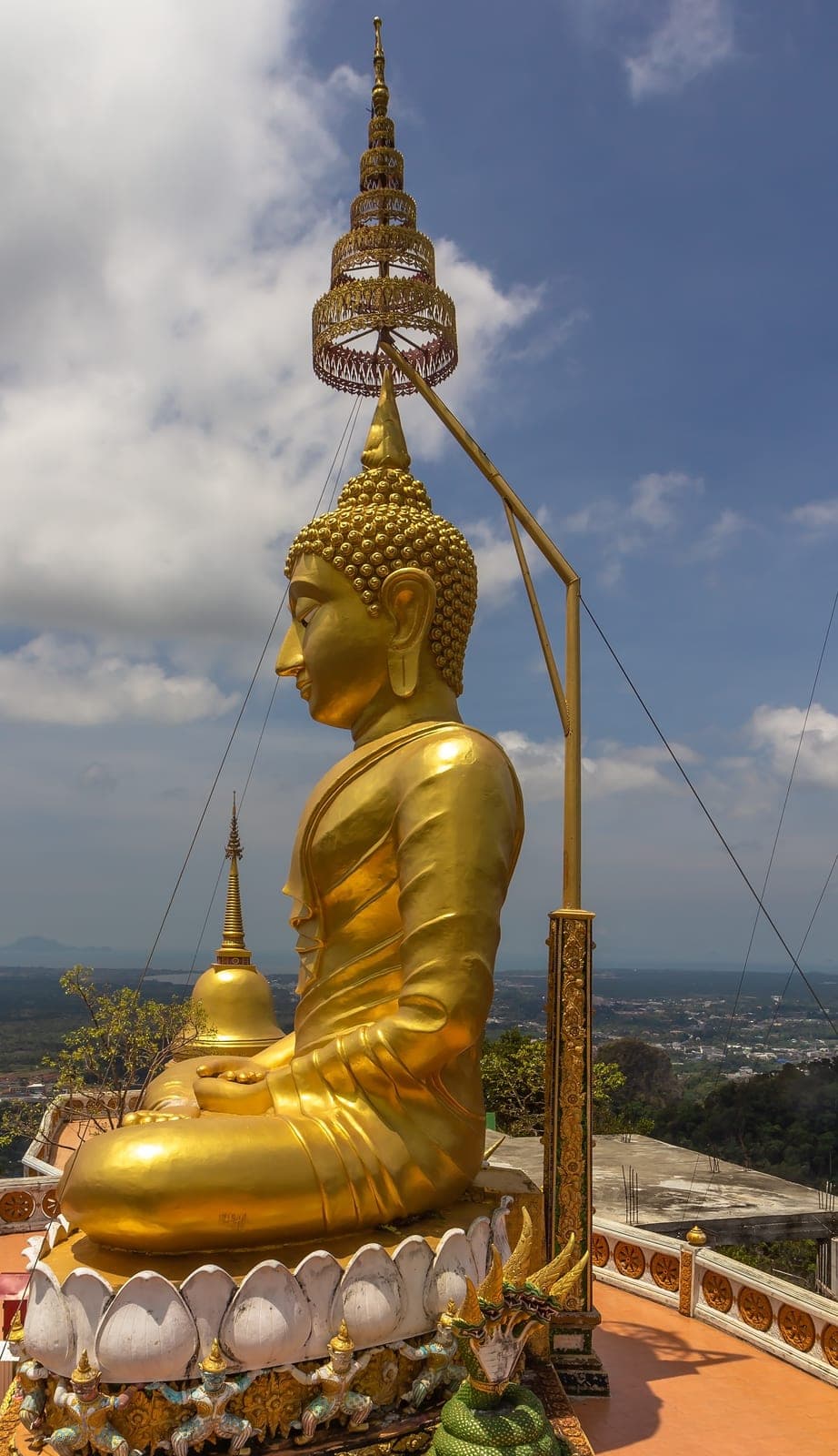The seated golden Buddha at the top of Tiger Cave Temple in profile, a gilded stupa beside it and the plain far below