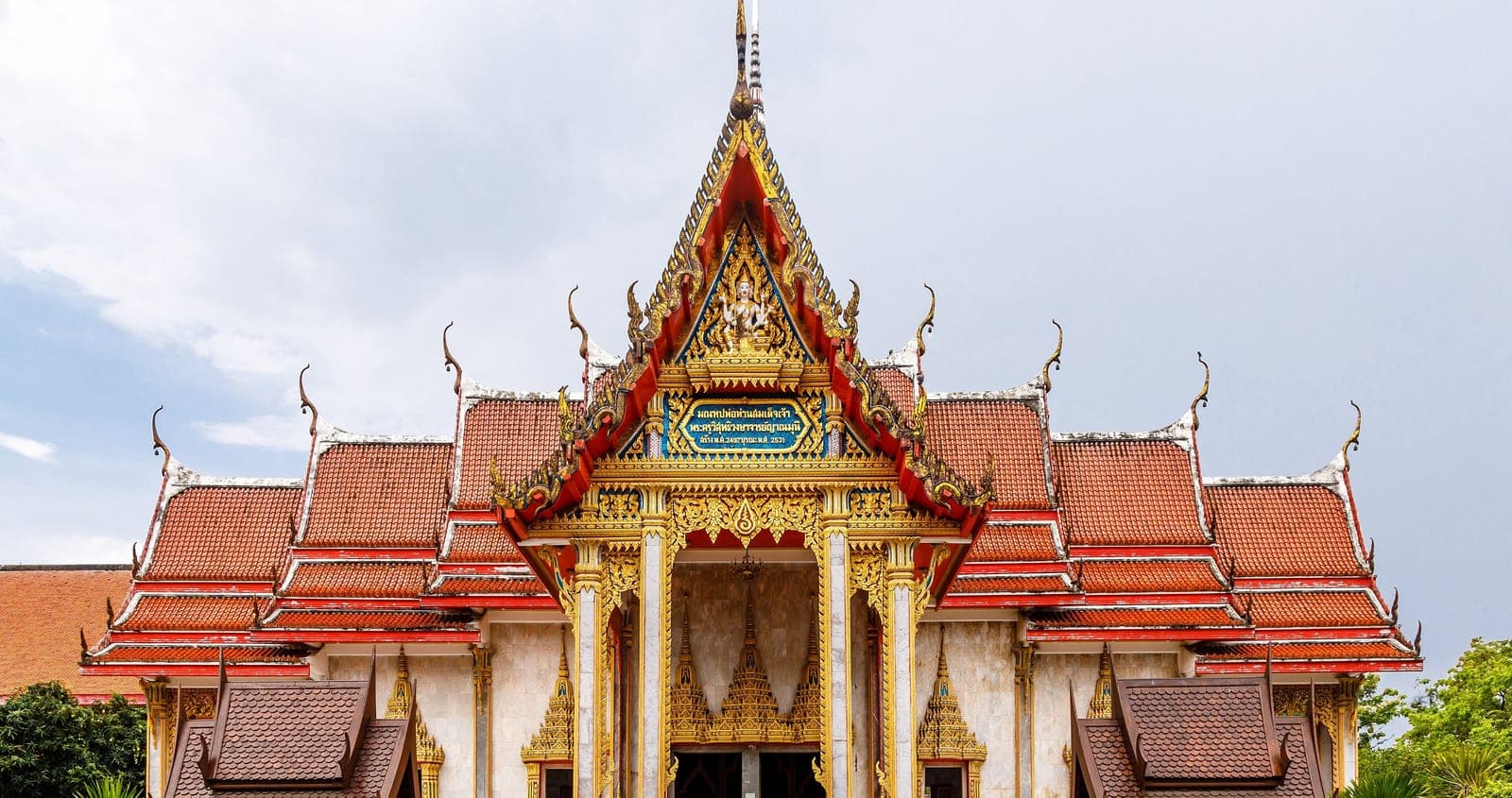 The front of a temple hall with tiered red-tiled roofs, a gilded gable and gold-trimmed columns under a grey sky
