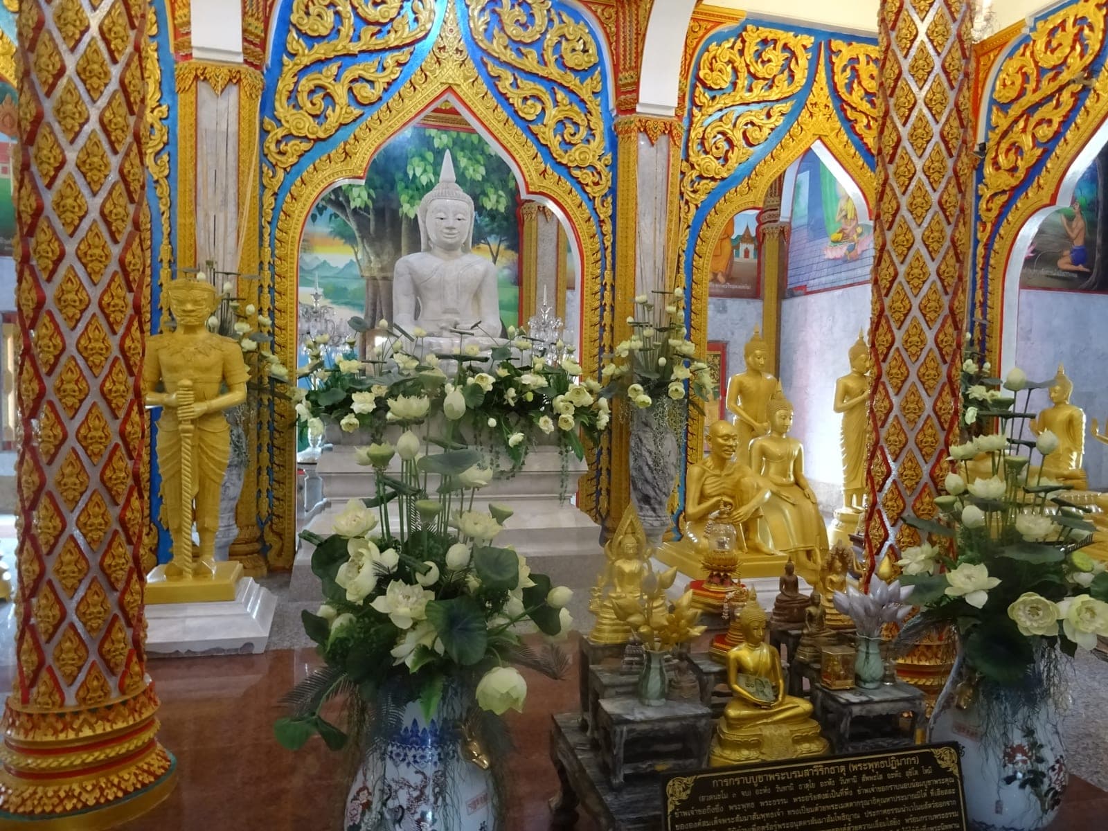 Inside the Grand Pagoda: a white Buddha image under a gilded blue arch, golden statues and white lotus flowers in vases, murals on the walls