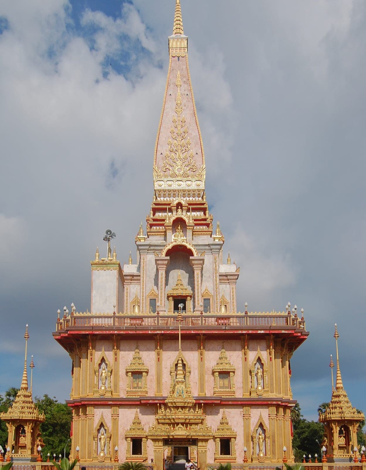 The Grand Pagoda of Wat Chalong, a tall pink and gold spire rising over a red-roofed base, small gilded pavilions either side under a cloudy blue sky