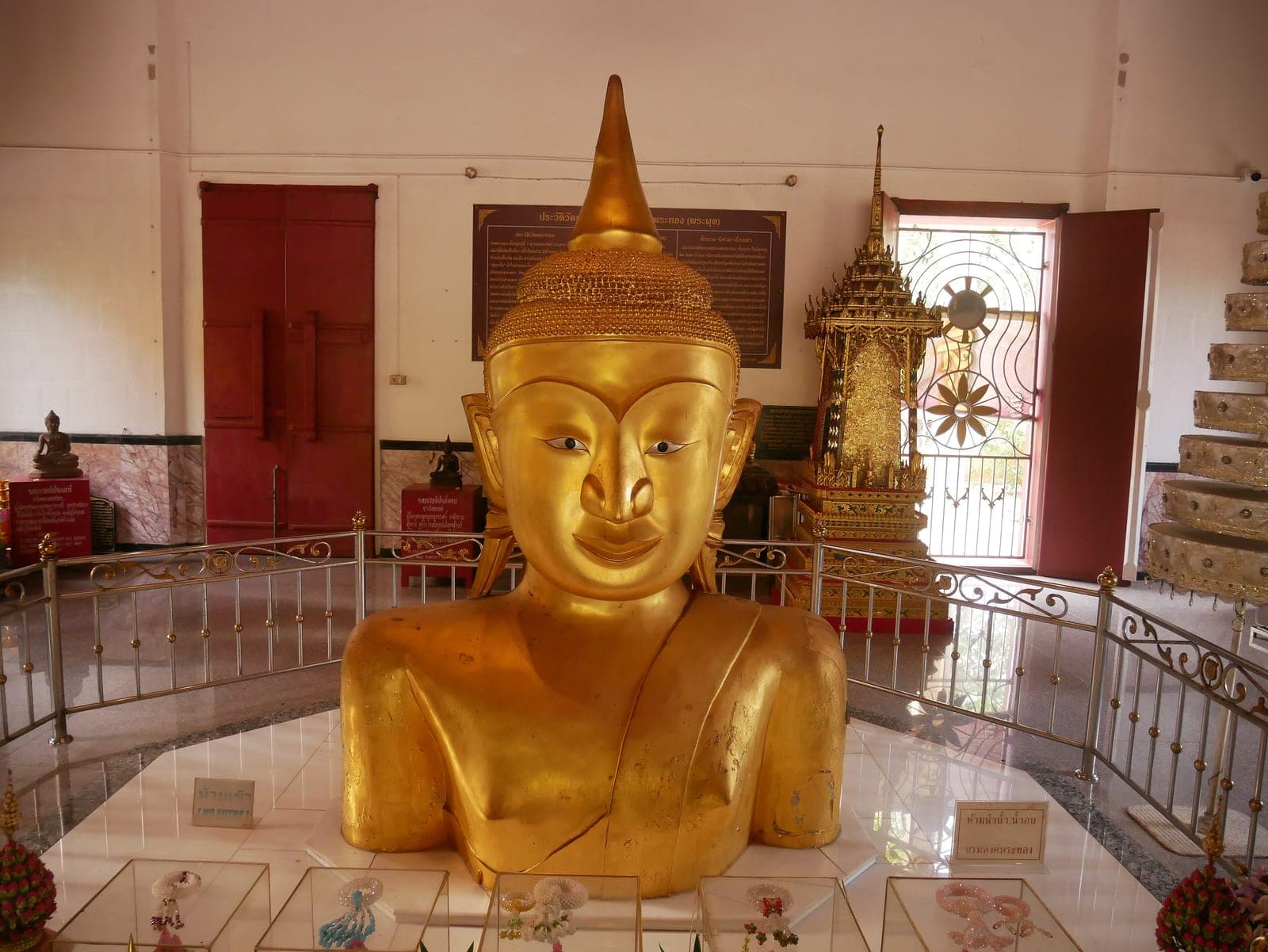The gilded head and shoulders of a Buddha image rising from a polished floor inside a temple hall, a railing around it and red doors behind