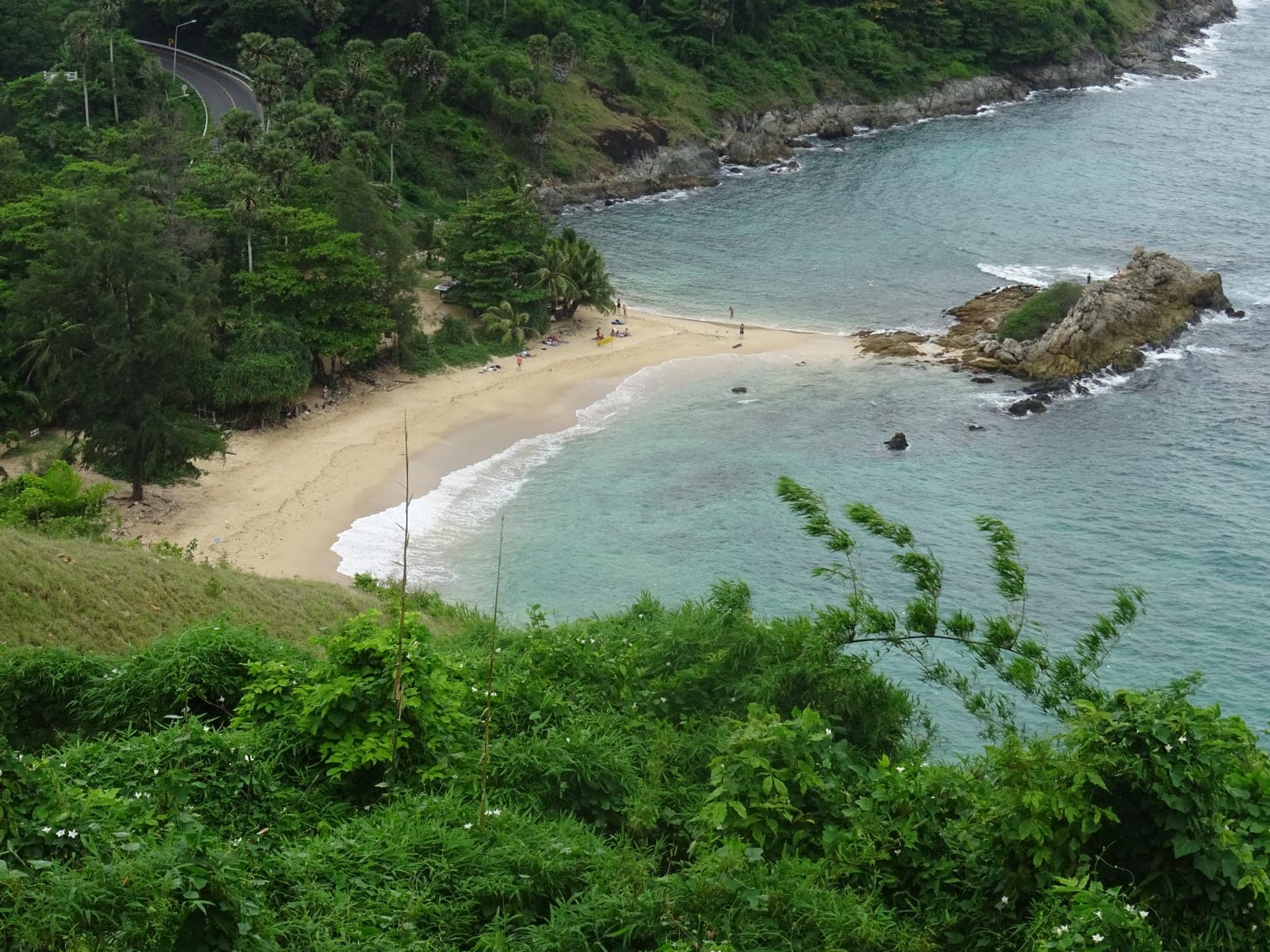 A small cove seen from above, a rocky point at one end of the sand and the coast road through the trees behind