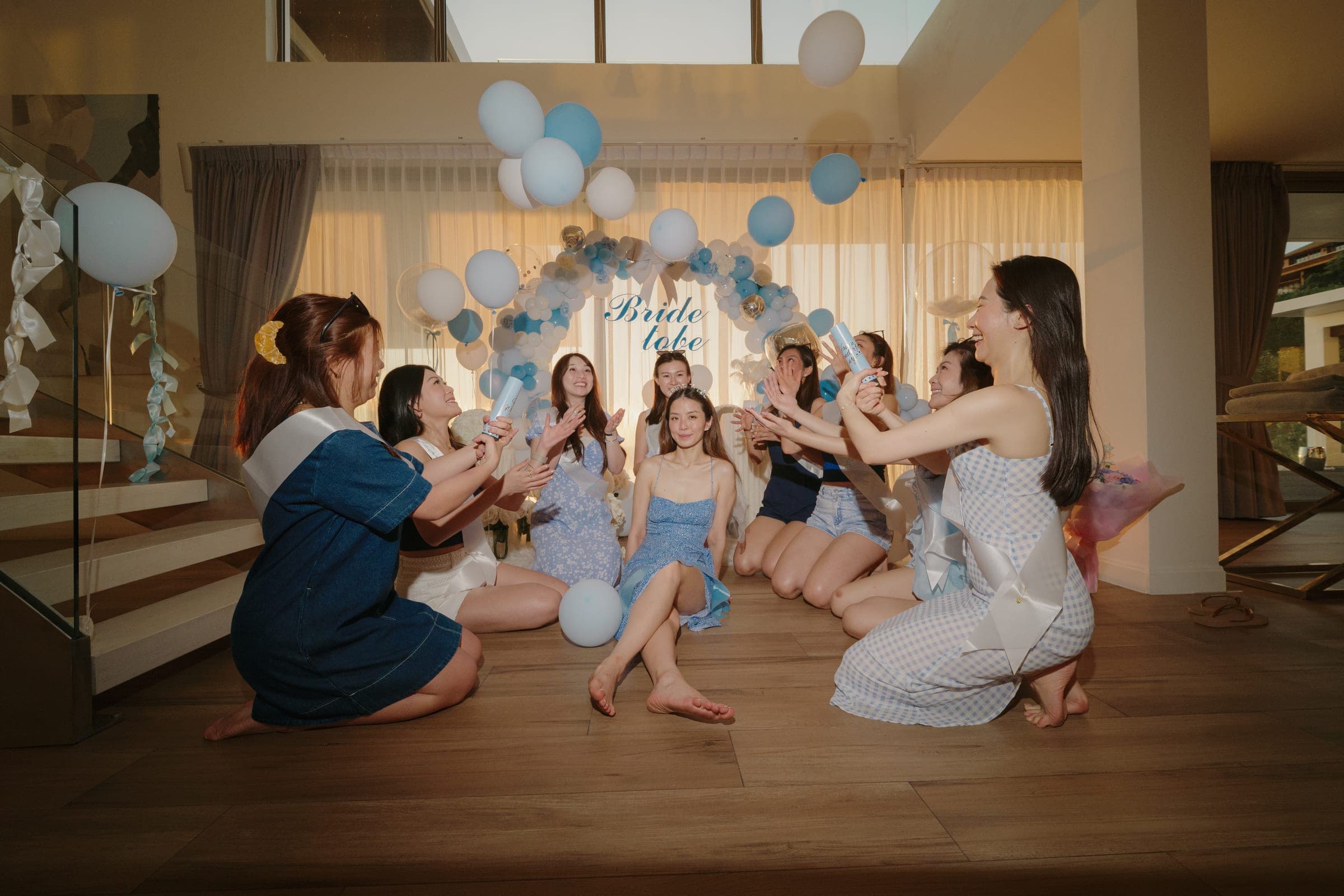 Friends in blue sitting on the floor round the bride-to-be under her balloon arch