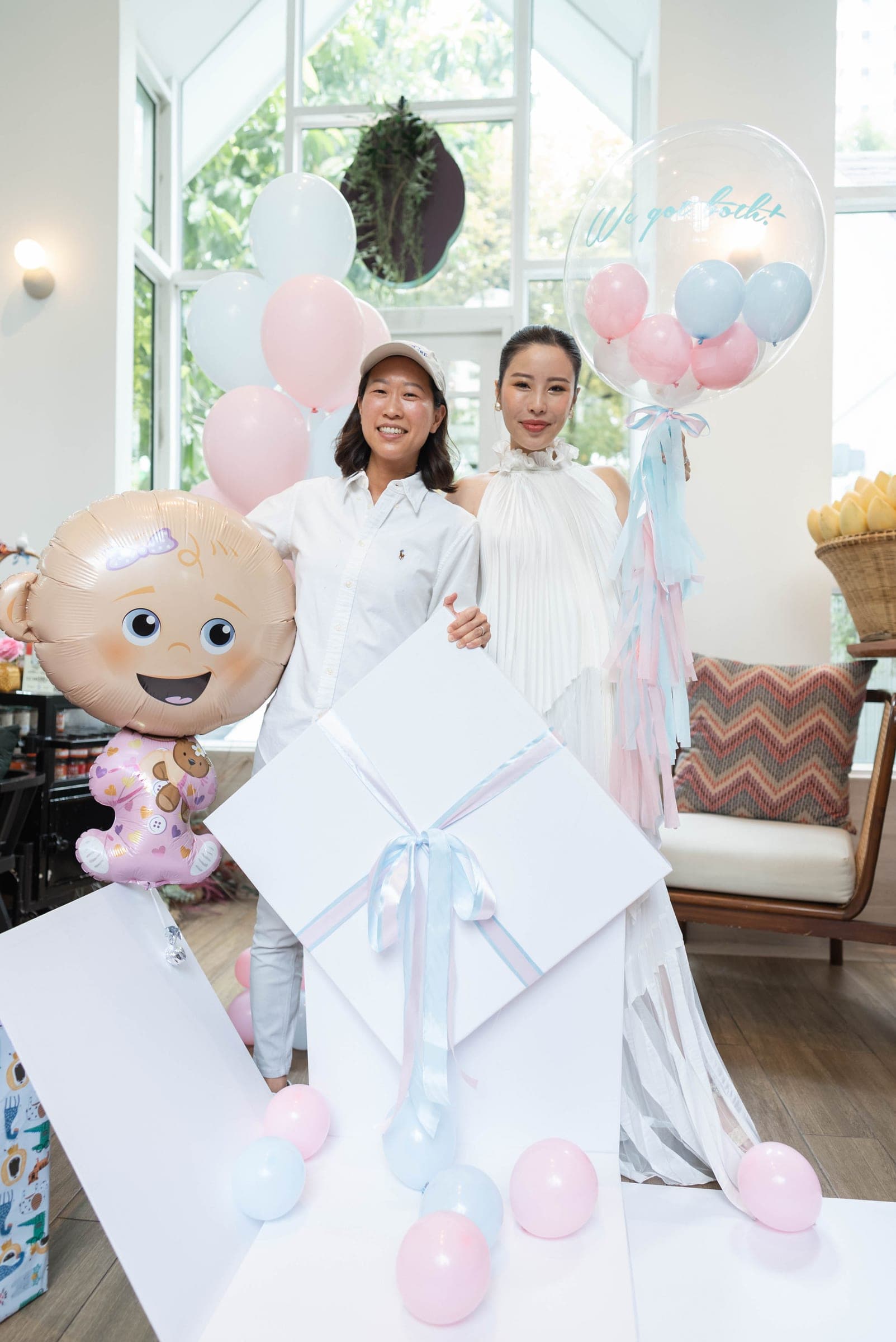 Mother-to-be and someone in a white cap with a giant baby balloon and a gift box, pink and blue balloons all around