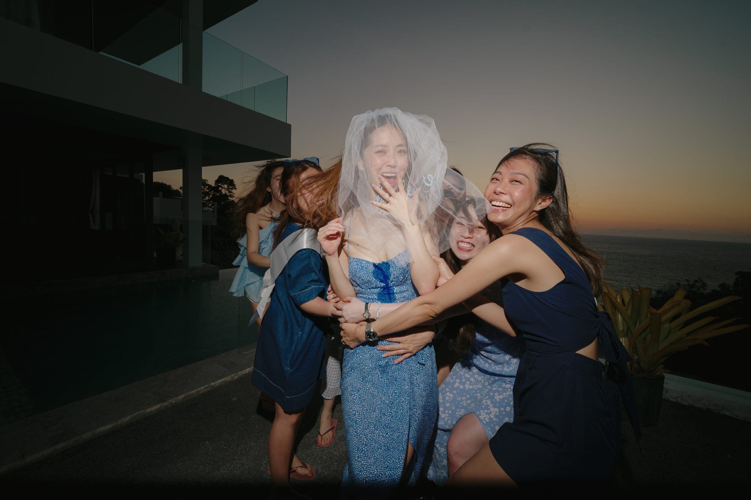Bachelorette group in blue laughing around the bride-to-be in a veil beside a villa pool at sunset