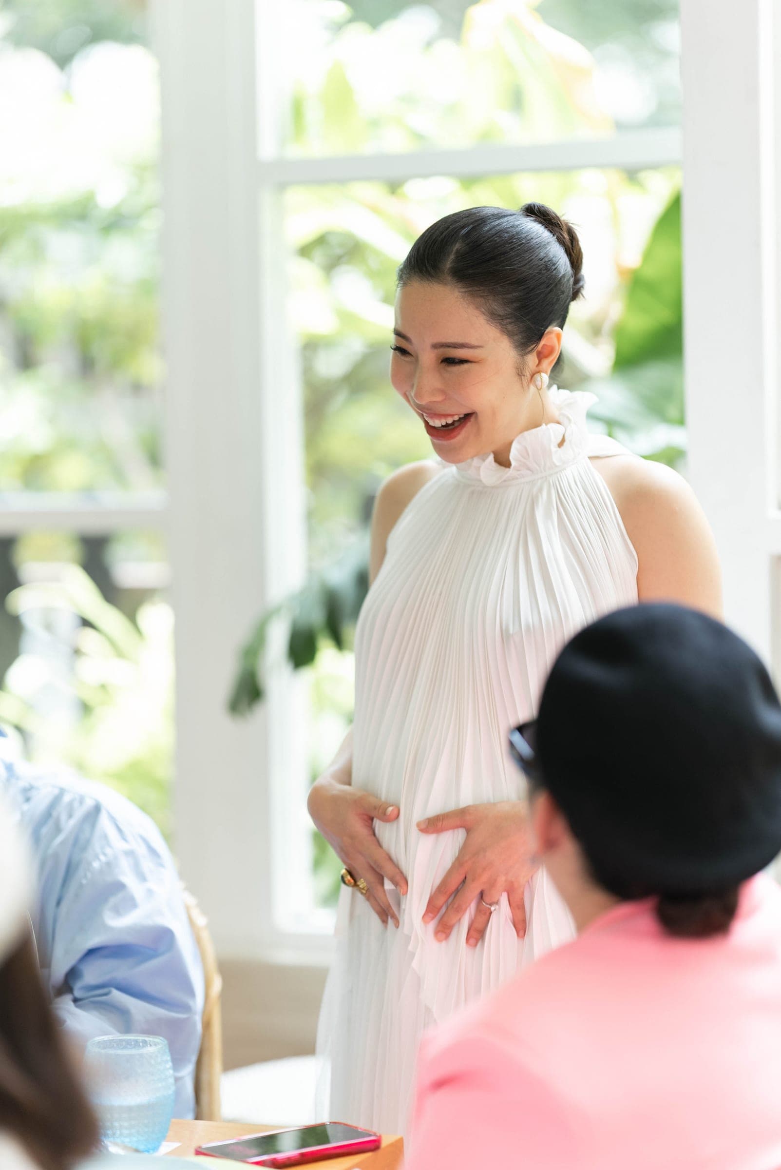 Mother-to-be in a white pleated dress laughing with guests at the table