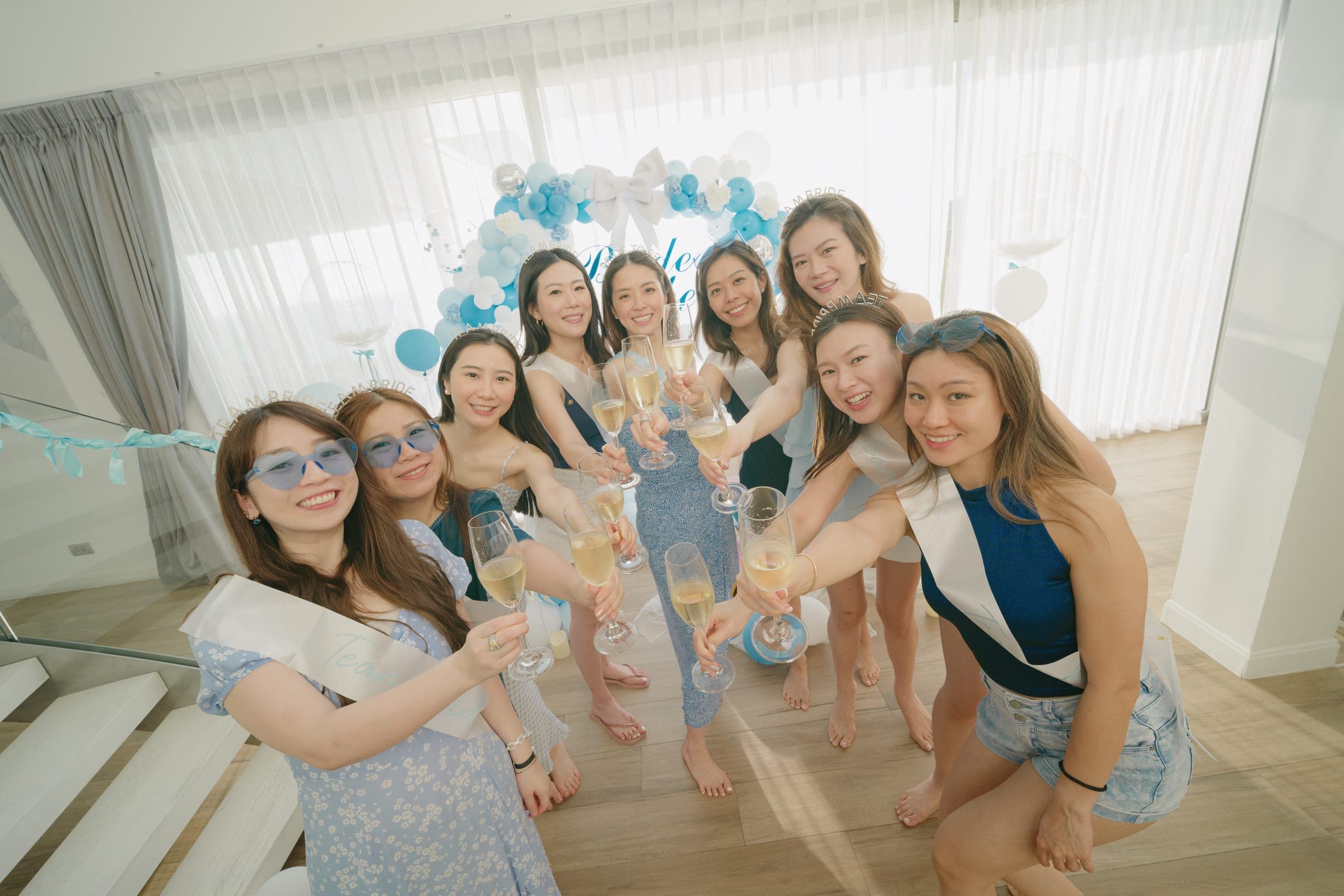 Friends in blue raising champagne flutes in front of a balloon arch for the bride-to-be in a bright villa