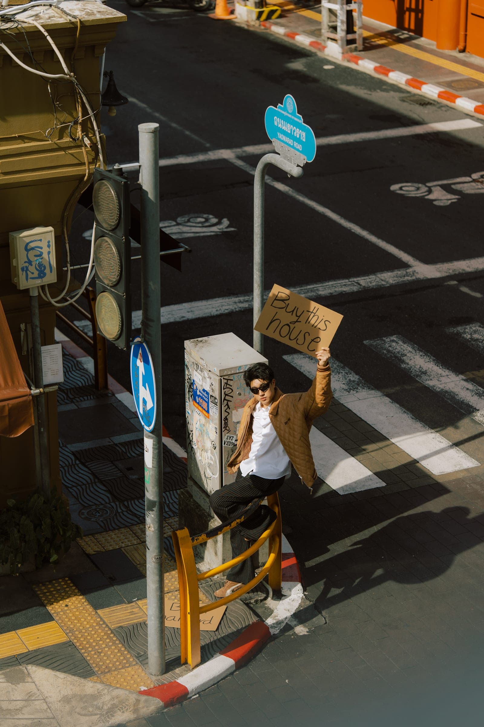 Man in sunglasses and a tan quilted jacket perched on a yellow railing at an Old Town street corner, holding up a hand-lettered cardboard sign