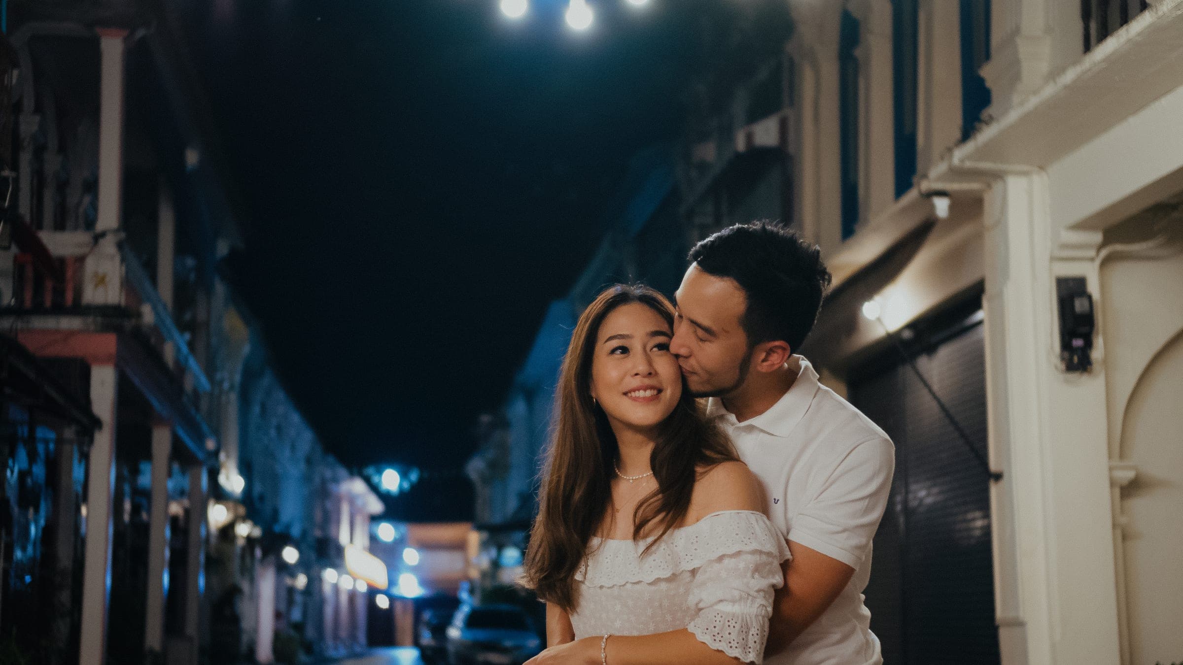 Man kissing his partner’s cheek from behind as she smiles, an Old Town lane lit up at night behind them