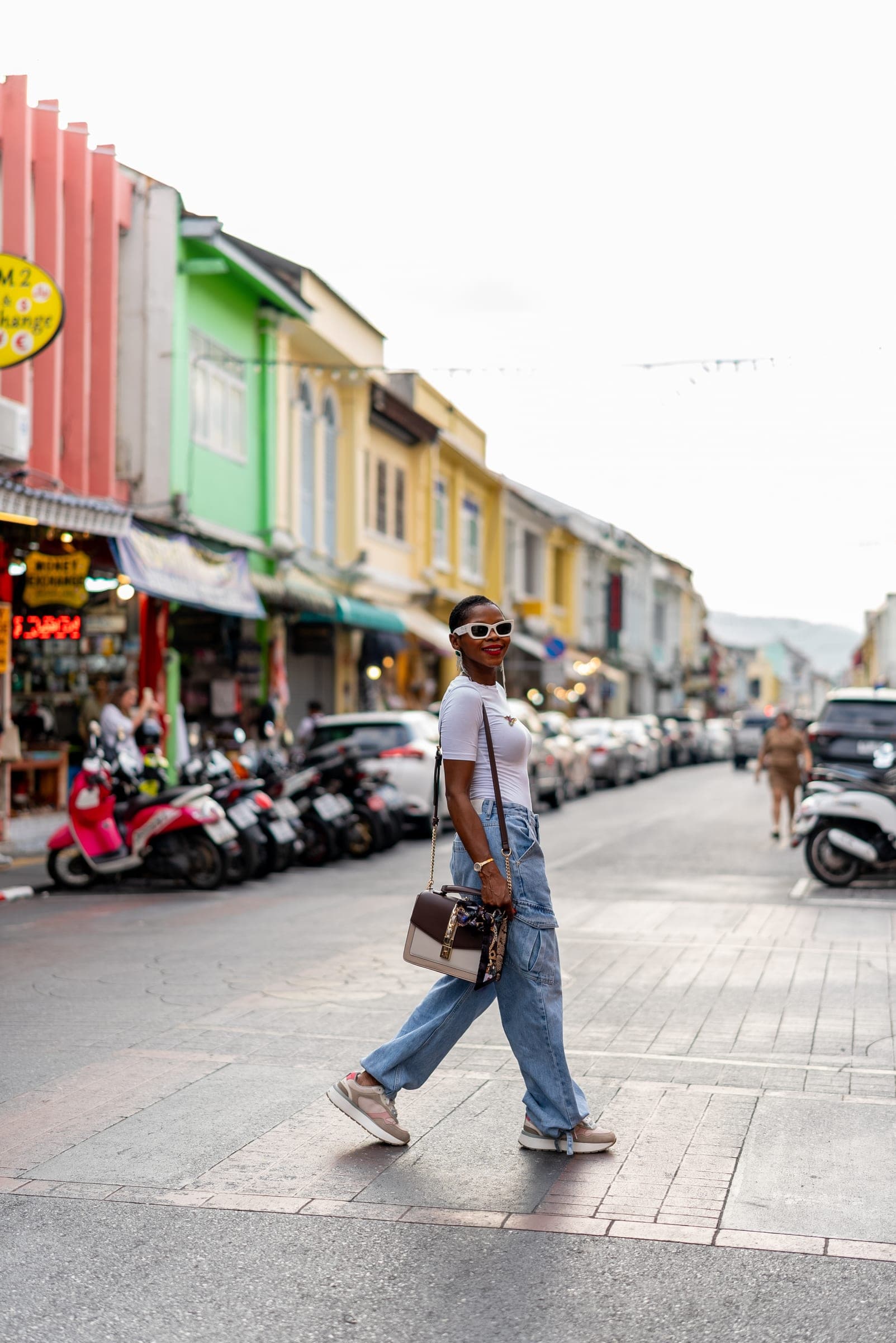Woman in white sunglasses and wide-leg jeans crossing an Old Town street, pink, green and yellow shophouses behind