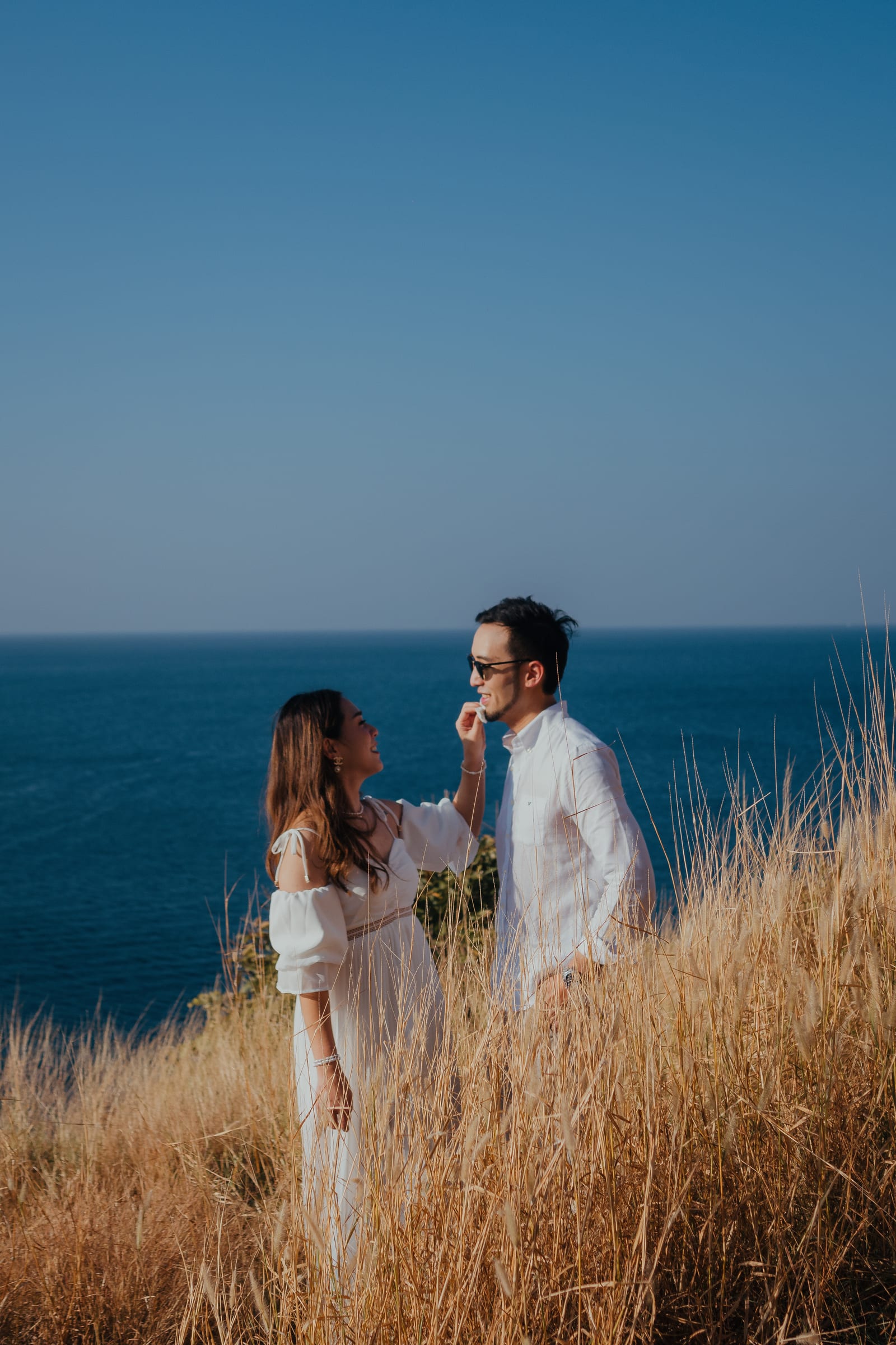 Couple in white on a grassy cliff, she straightening his collar, deep blue sea behind
