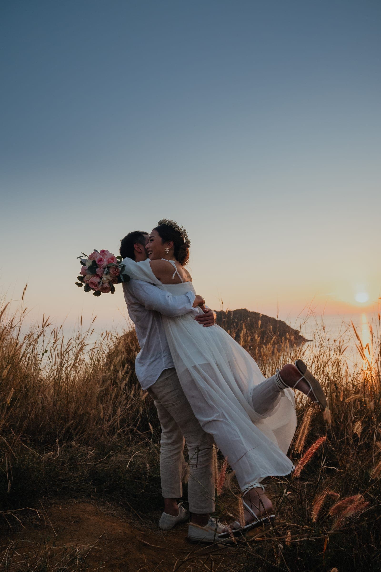 Man lifting his partner off her feet on a grassy headland as the sun sets over the sea behind them