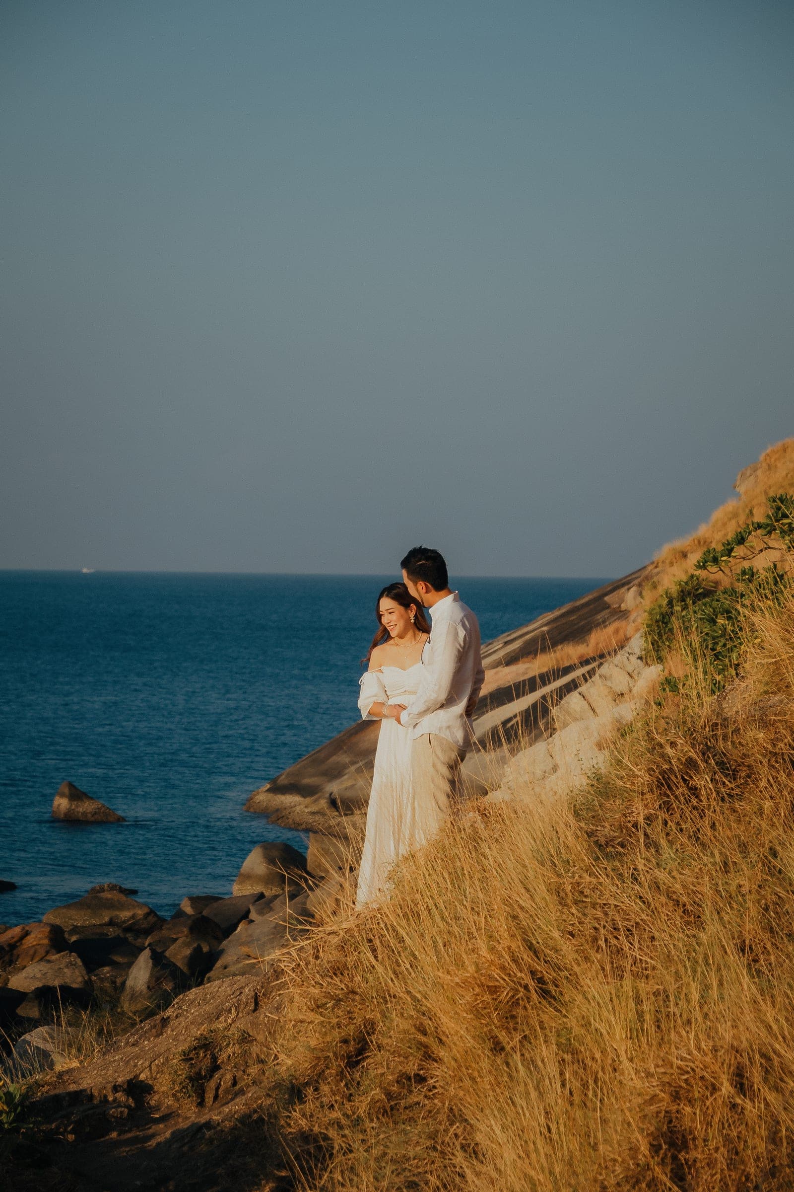 Couple in white standing in the long grass above the rocks and the dark blue sea, her dress bright against the cliff