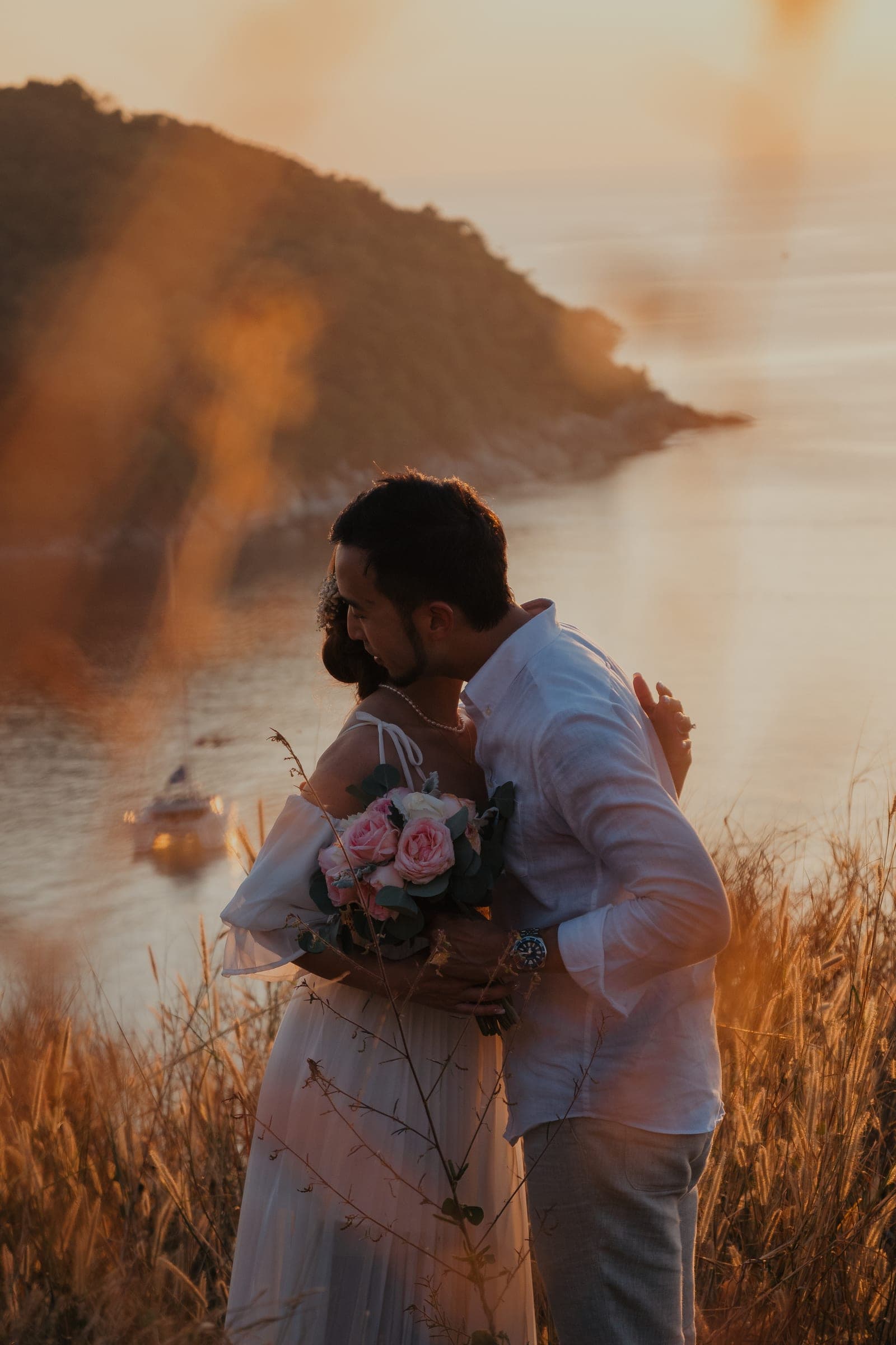 Couple holding each other in tall dry grass above the sea, a bouquet between them, the low sun flaring gold across the frame