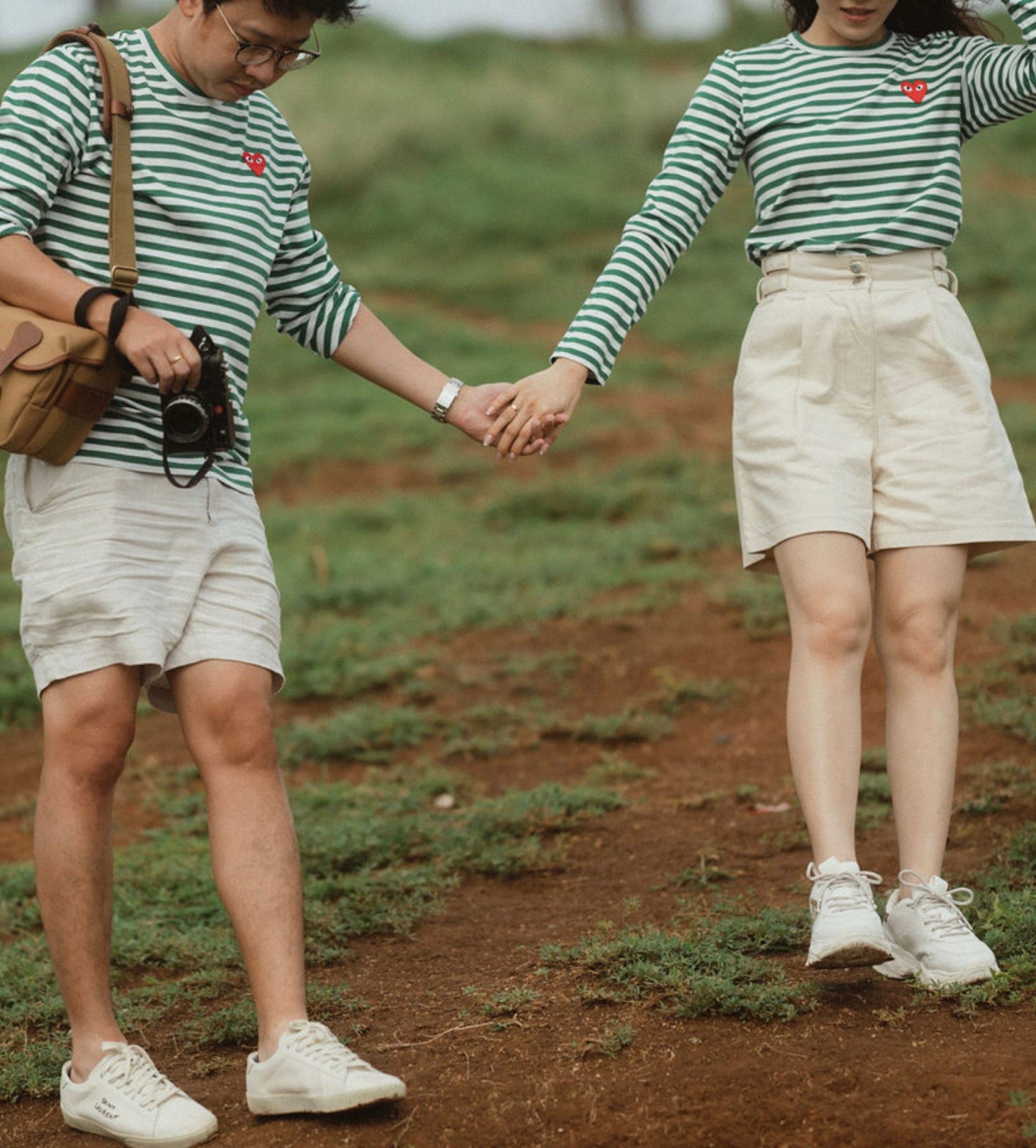 Couple in matching green-striped tops holding hands as they walk across the grass, one carrying a camera