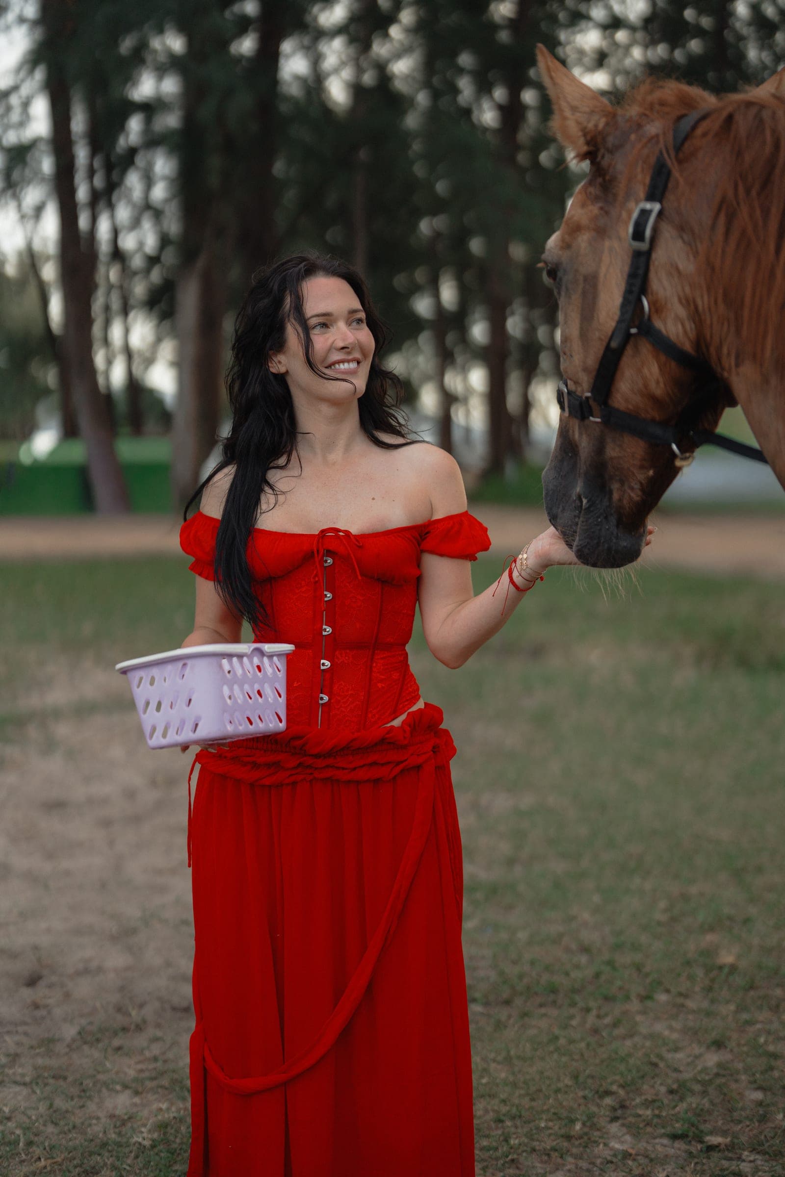 Woman in a red off-the-shoulder dress laughing as she holds a basket out to a chestnut horse under casuarina trees