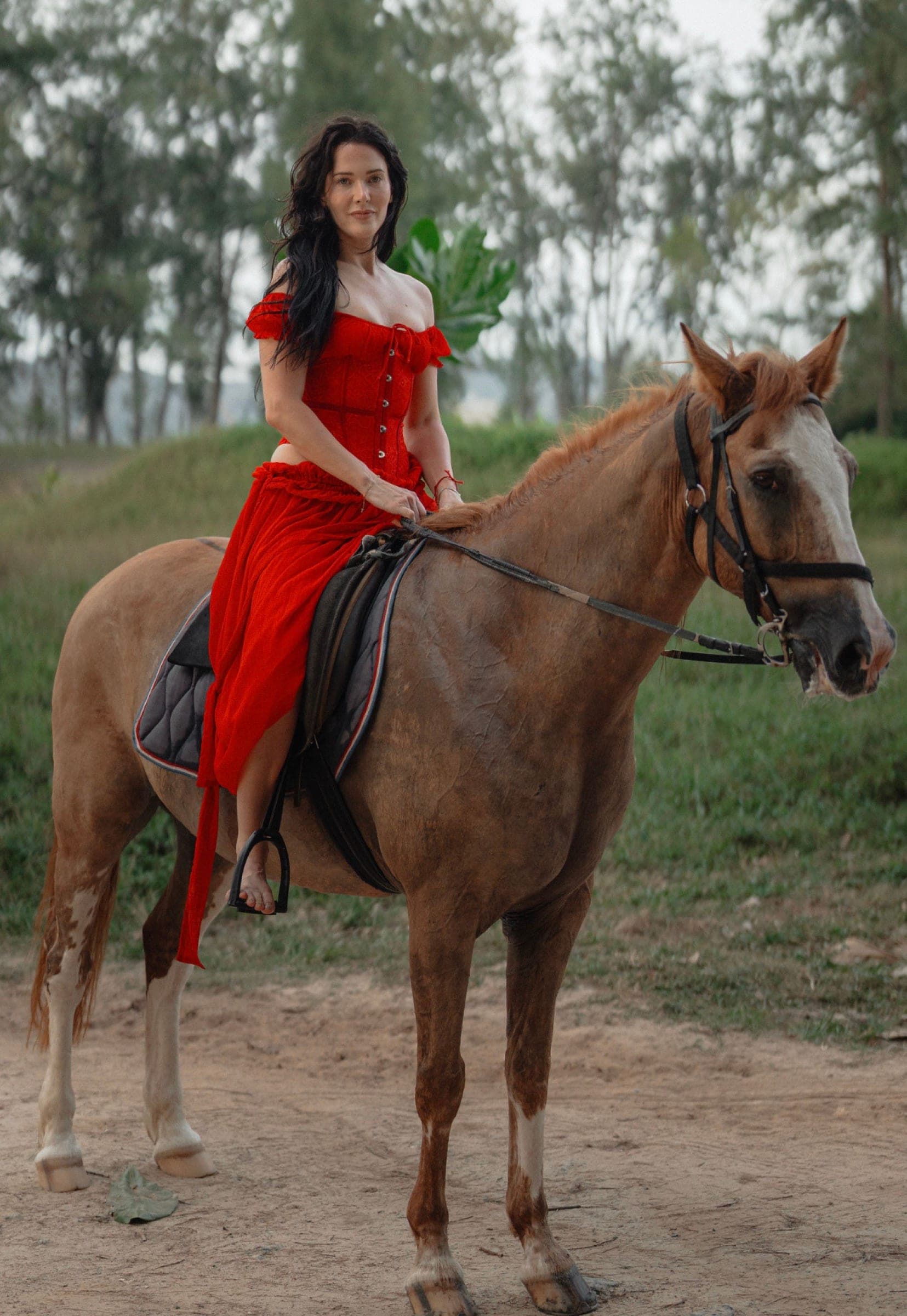 Woman in a red dress on a chestnut horse on a sandy track, casuarina trees behind
