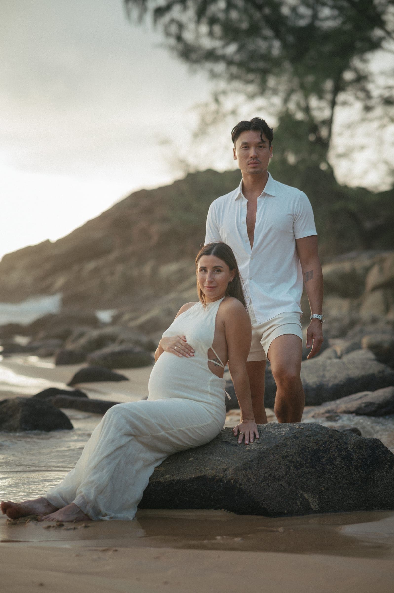 Couple on a rock at the beach, she sitting in a white dress with a hand on her bump, he standing beside her