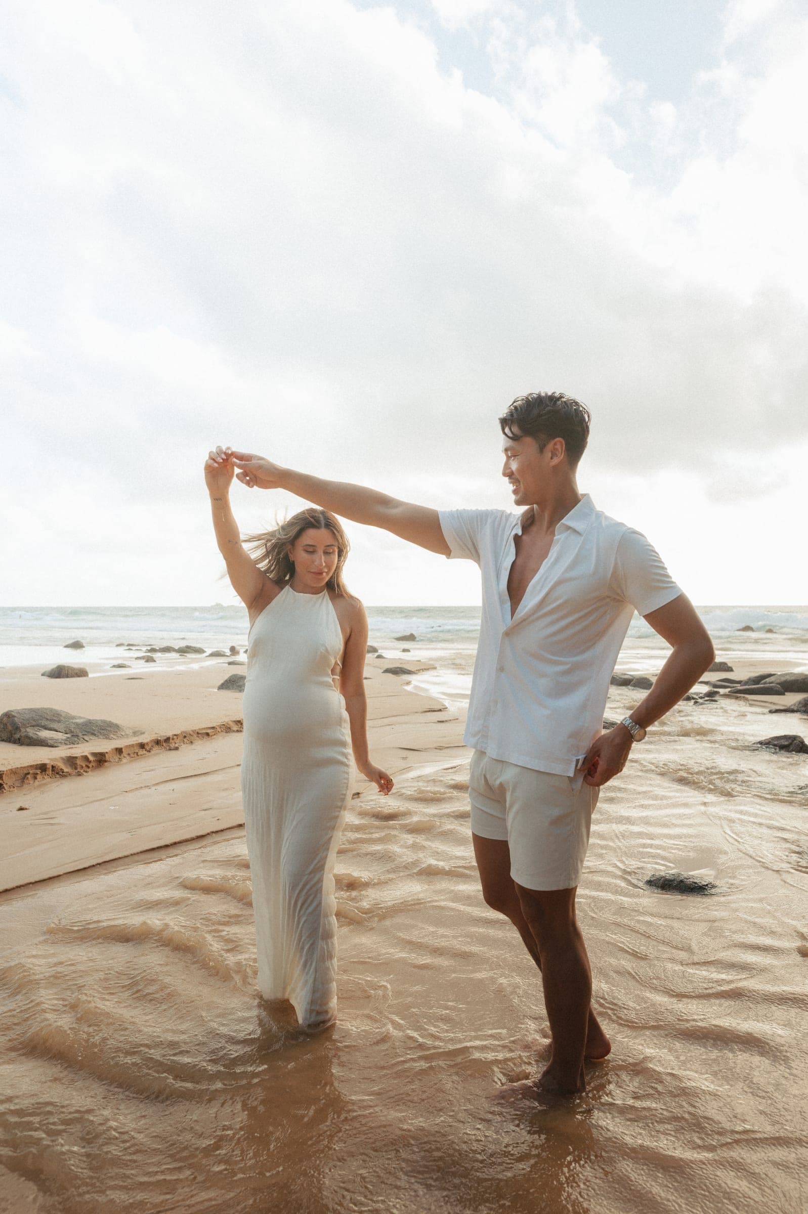 Man twirling his partner by the hand at the water’s edge, both in white, waves breaking behind