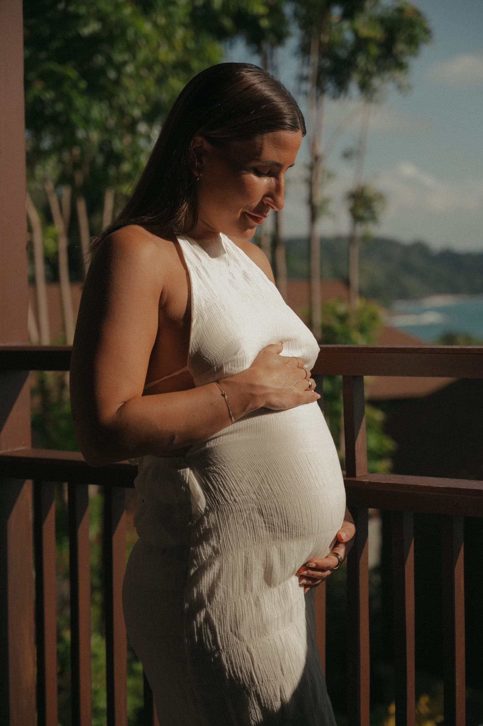 Expectant mother on a wooden balcony, hands on her bump, palms and the bay behind her