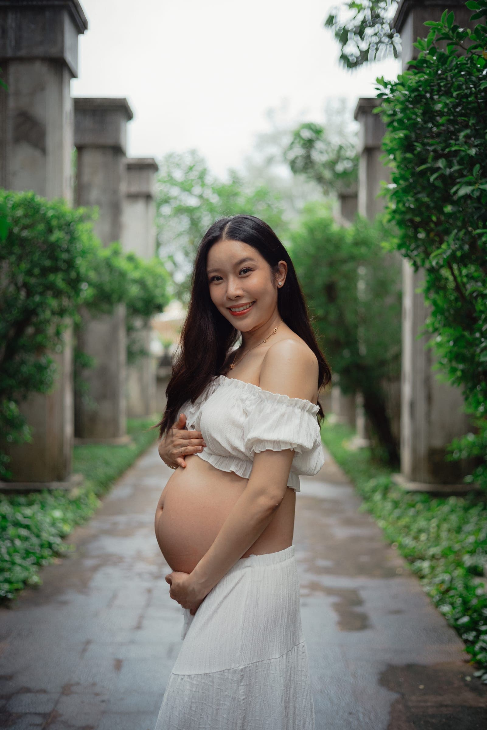 Expectant mother in a cream top and long white skirt standing on a wet garden path between stone columns and greenery