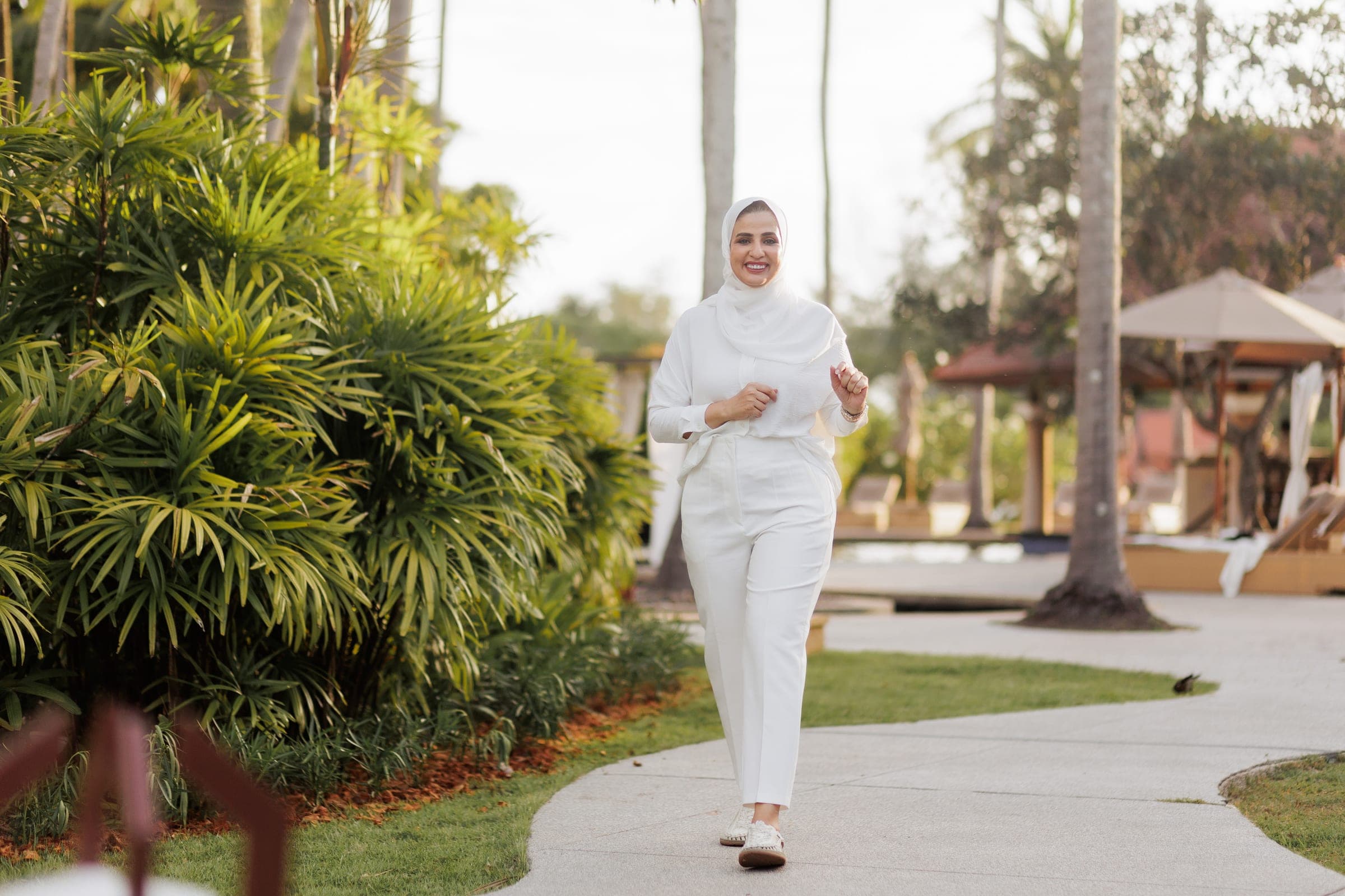 Woman in a white headscarf, blouse and trousers walking along a garden path, palms and sun loungers behind