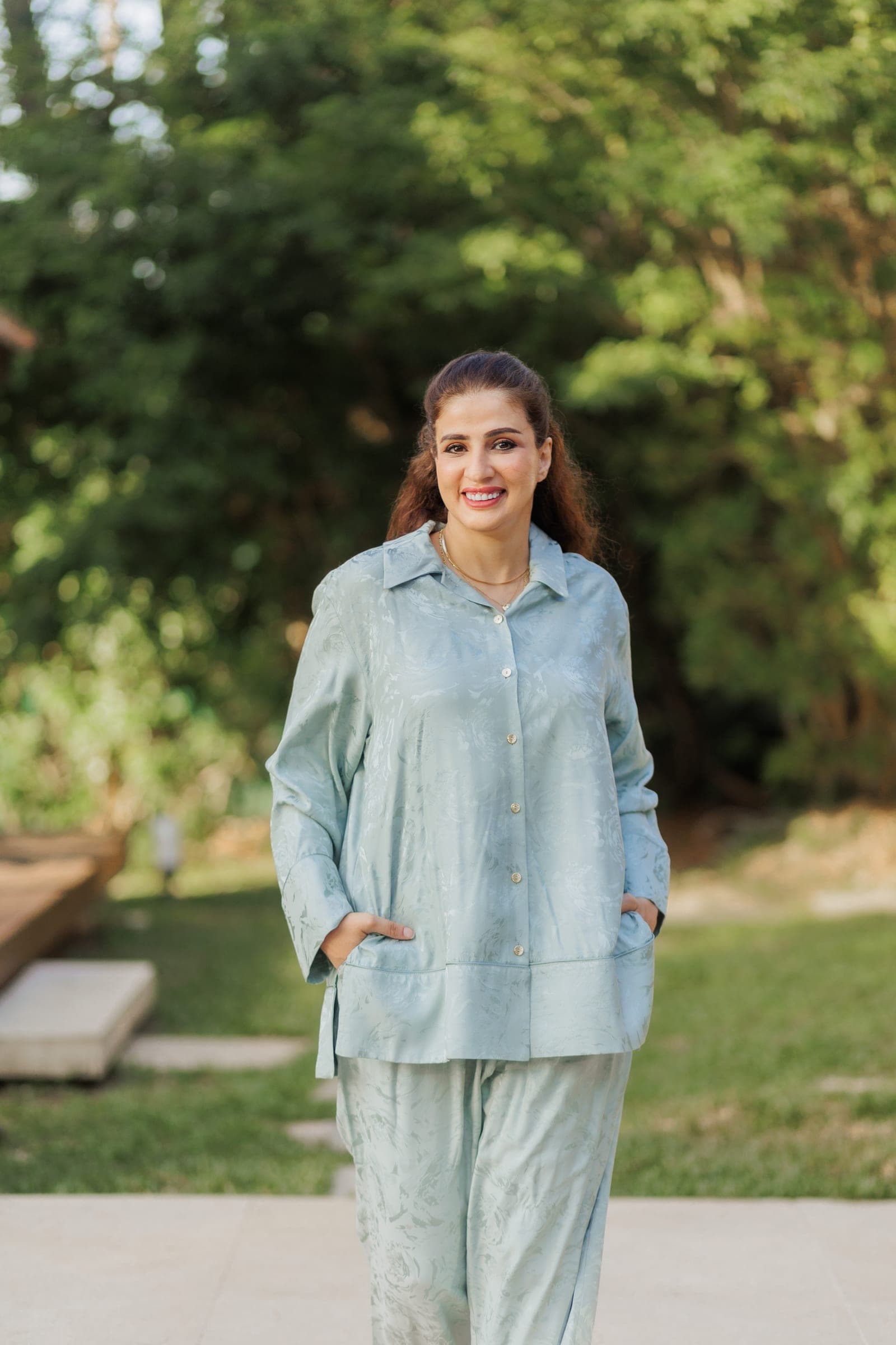 Smiling woman in a pale blue-green satin shirt and trousers, hands in her pockets, tall trees behind