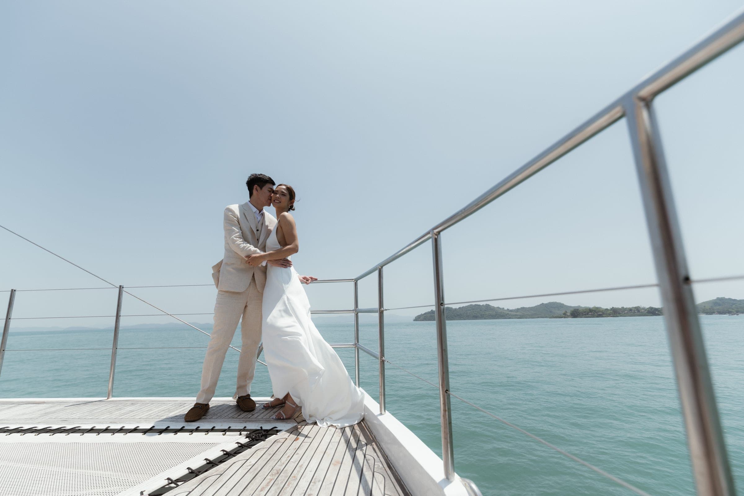 Couple in wedding white at the bow of a catamaran, islands scattered behind