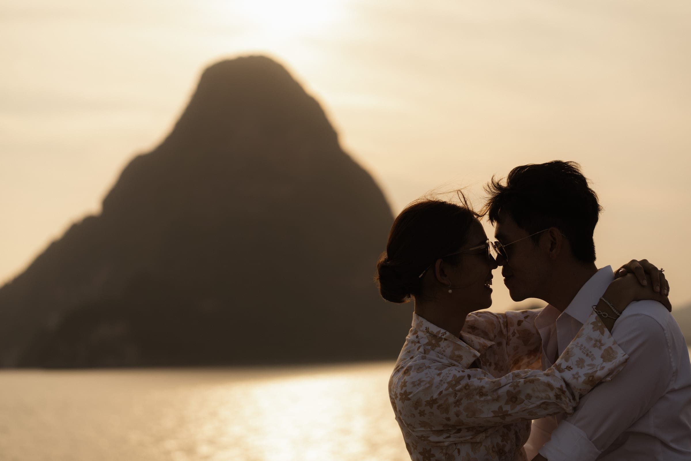 Couple silhouetted nose to nose against a gold sunset, a limestone island rising behind them